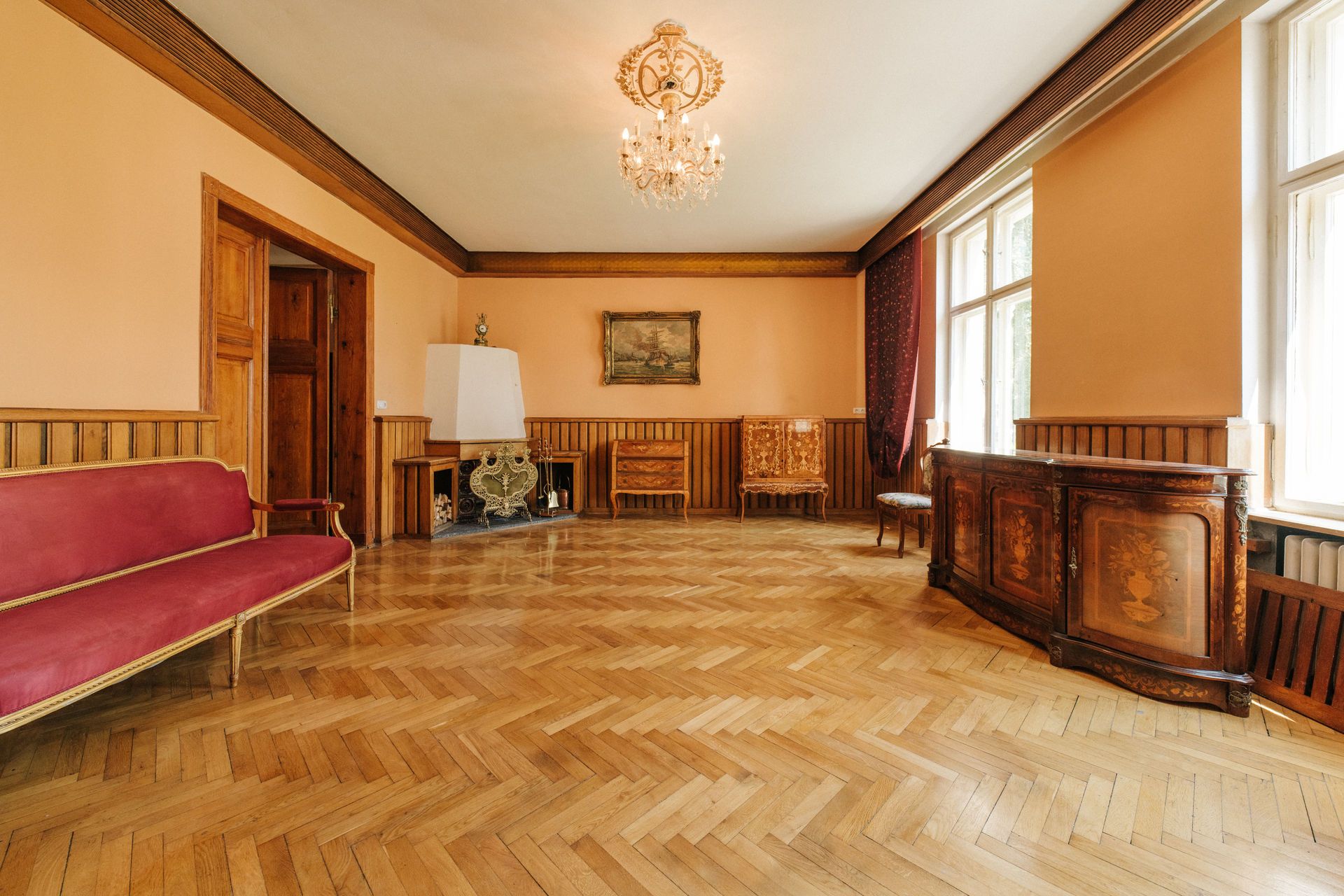 Geräumiges Zimmer mit Parkettboden, Kronleuchter, roten Sitzbänken und antiken Holzmöbeln.