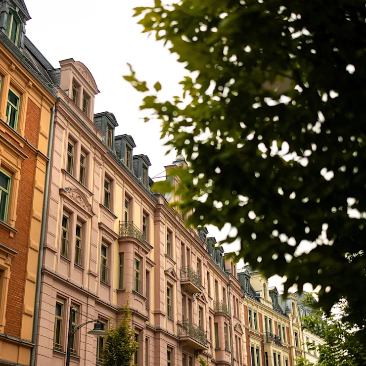 Mehrstöckige Wohnhäuser mit verzierten Fassaden und Balkonen, daneben ein Baum im Vordergrund.