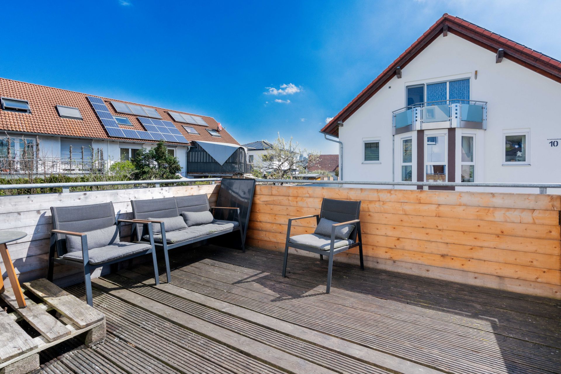 Sonnige Holzterrasse mit Sitzgelegenheiten, Blick auf Nachbarhäuser und blauen Himmel.
