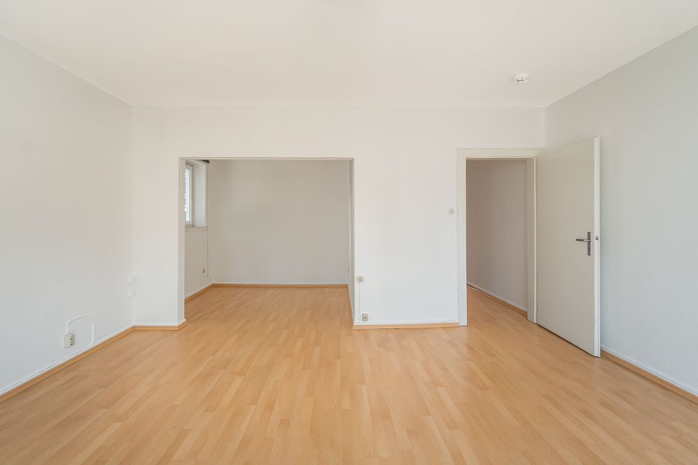 Leerer Raum mit hellem Laminatboden, weißen Wänden und zwei offenen Durchgängen.