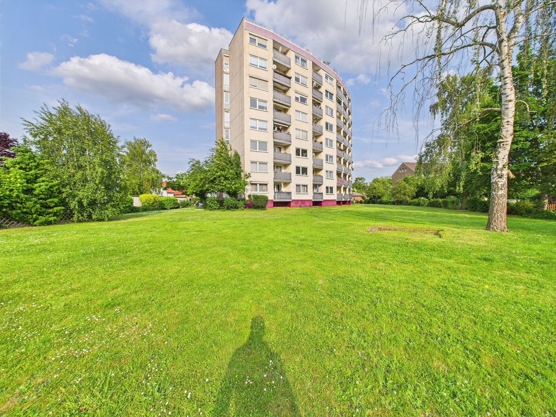 Mehrstöckiges Wohnhaus mit Balkonen, umgeben von einer großen grünen Wiese und Bäumen.