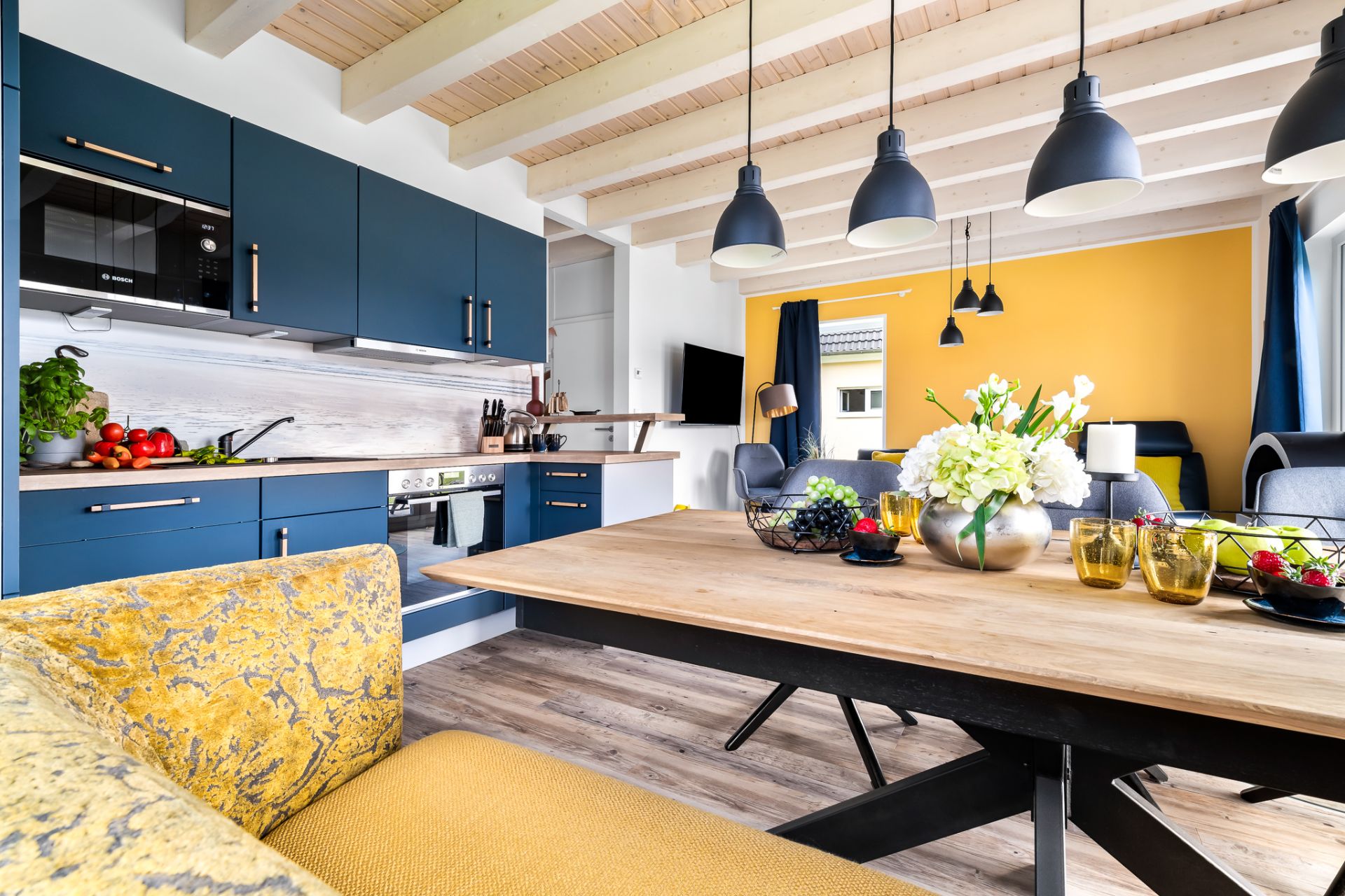 Moderne Wohnküche mit Holztisch, blauen Schränken und gelber Wand. Blumen und Obst auf dem Tisch.