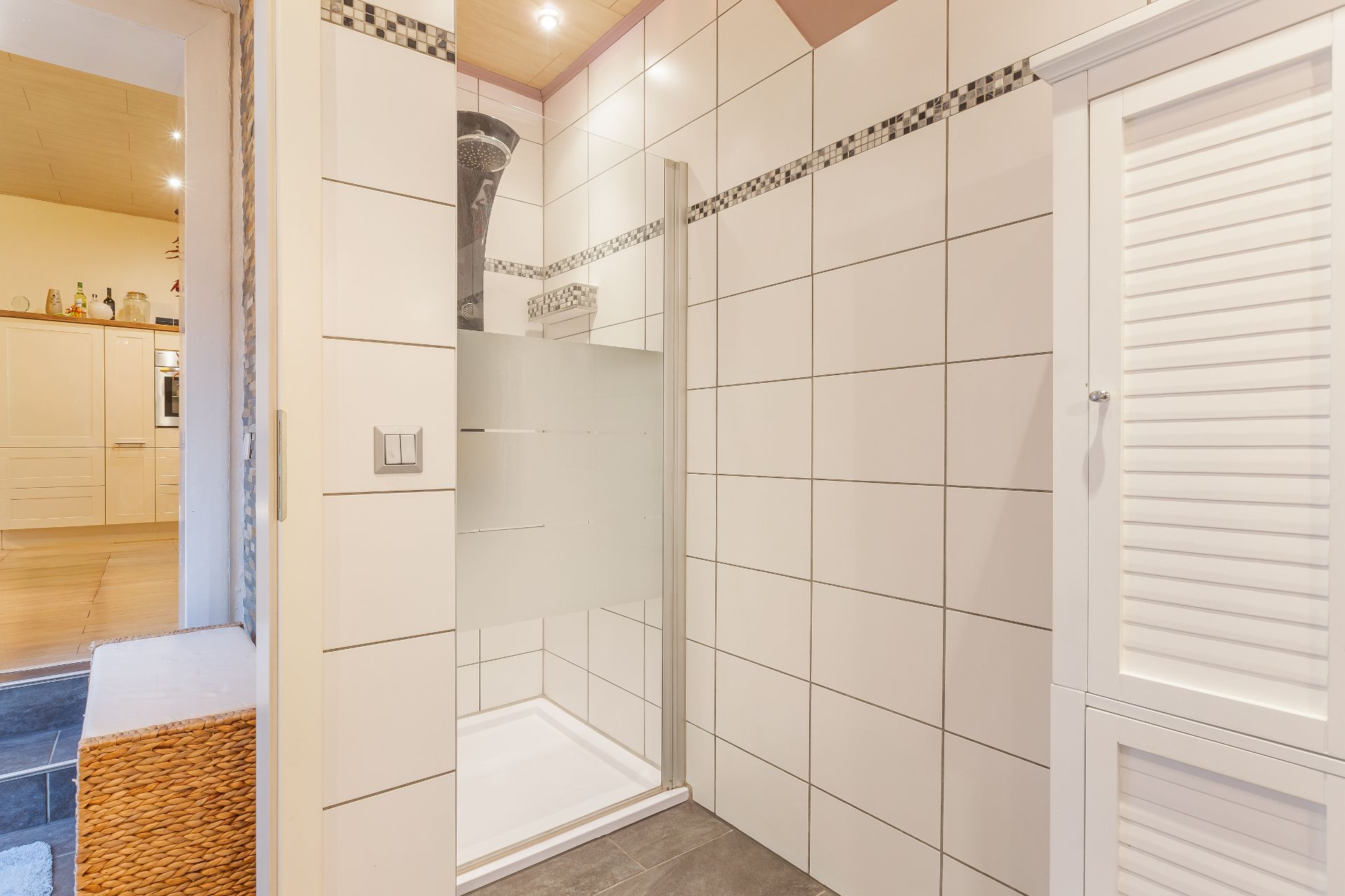Gefliestes Badezimmer mit Dusche, daneben ein weißer Schrank und ein Teil der Küche sichtbar.