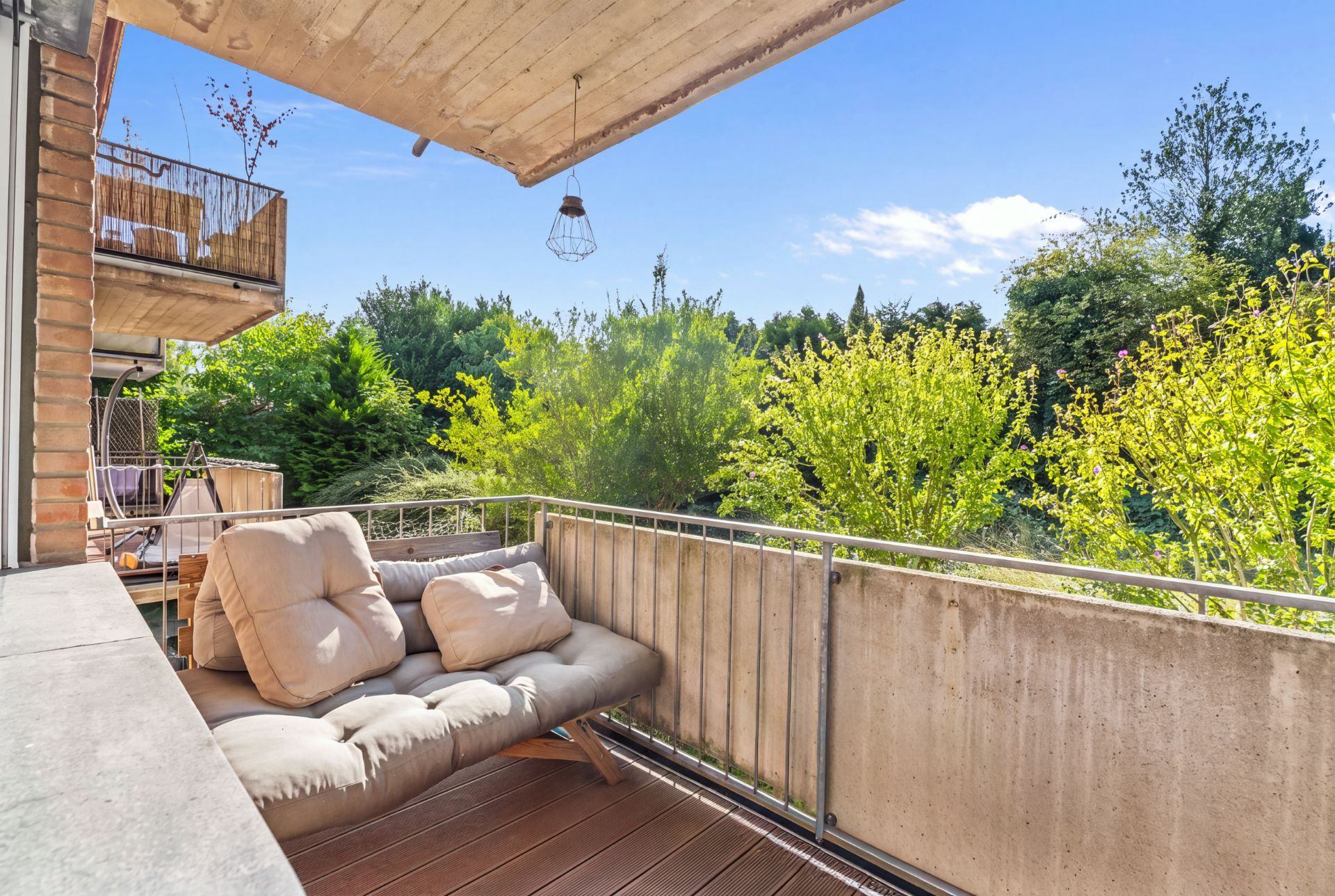 Balkon mit gemütlichem Sofa und Blick auf grüne Bäume an einem sonnigen Tag.