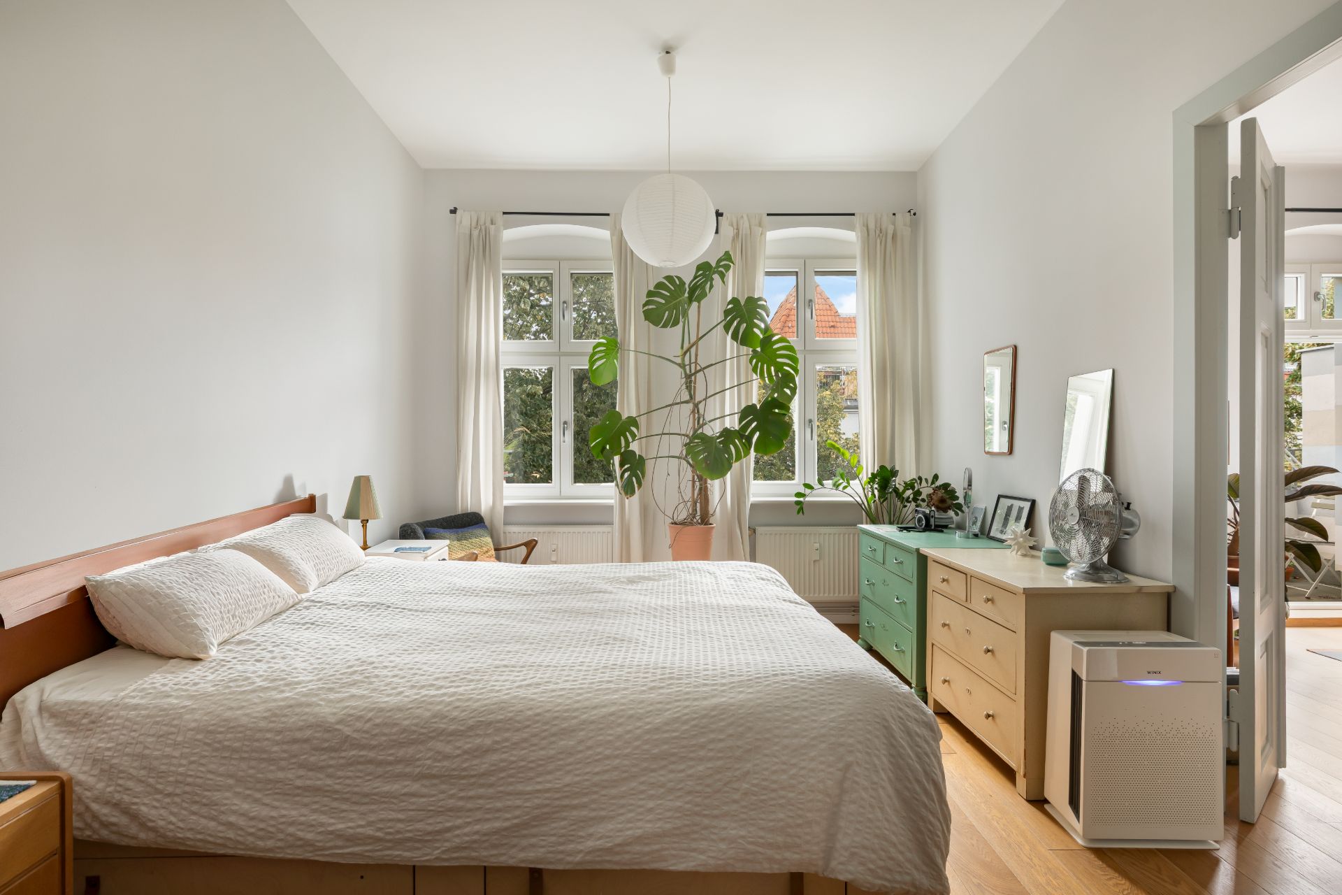 Helles Schlafzimmer mit Doppelbett, Pflanze am Fenster, Kommoden und Holzfußboden.