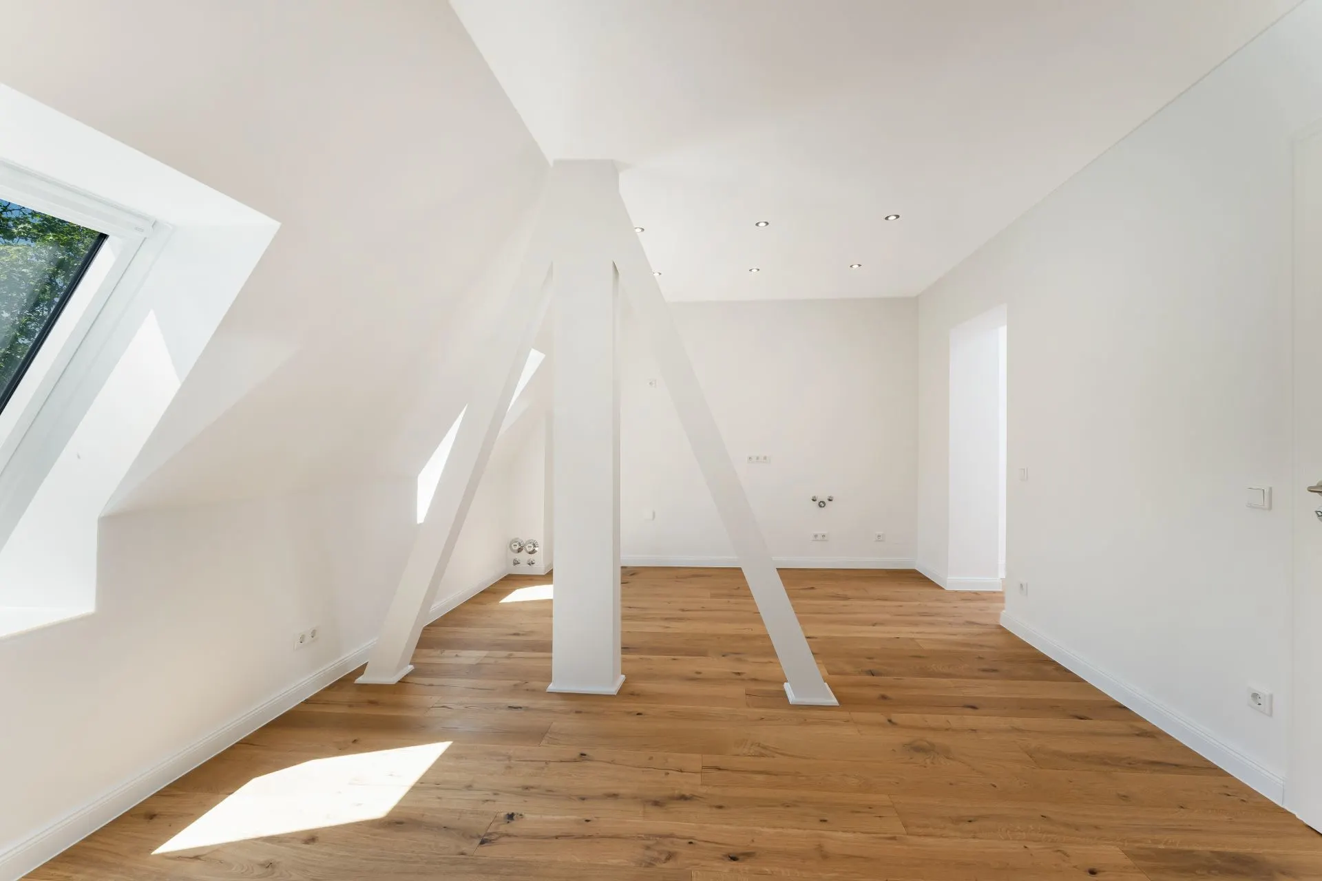 Heller Raum mit schräger Decke, großem Fenster, Holzboden und weißen Balken in der Mitte.