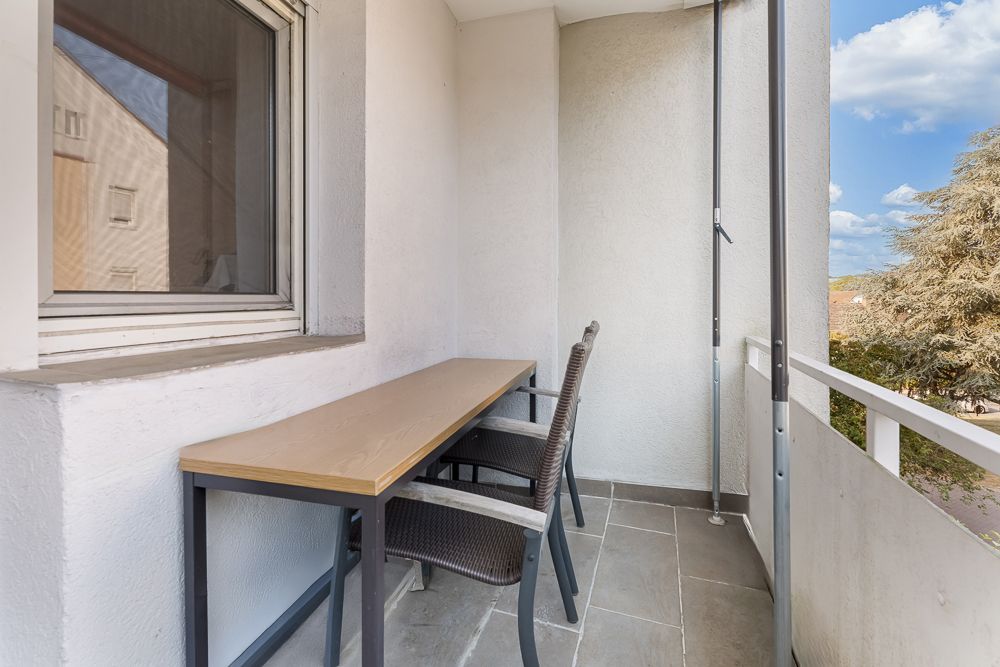 Überdachter Balkon mit zwei Stühlen und einem langen Tisch, Blick auf Bäume und blauen Himmel.
