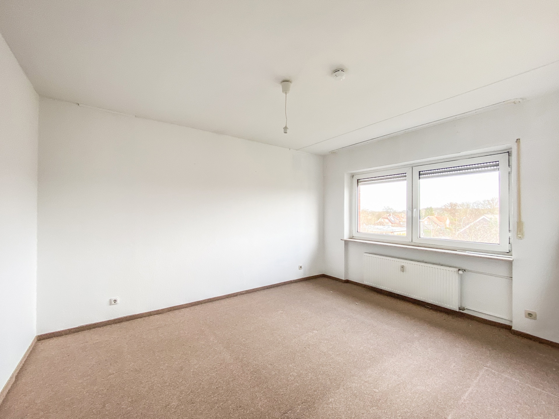 Leeres Zimmer mit Teppichboden, weißen Wänden und großem Fenster mit Blick nach draußen.