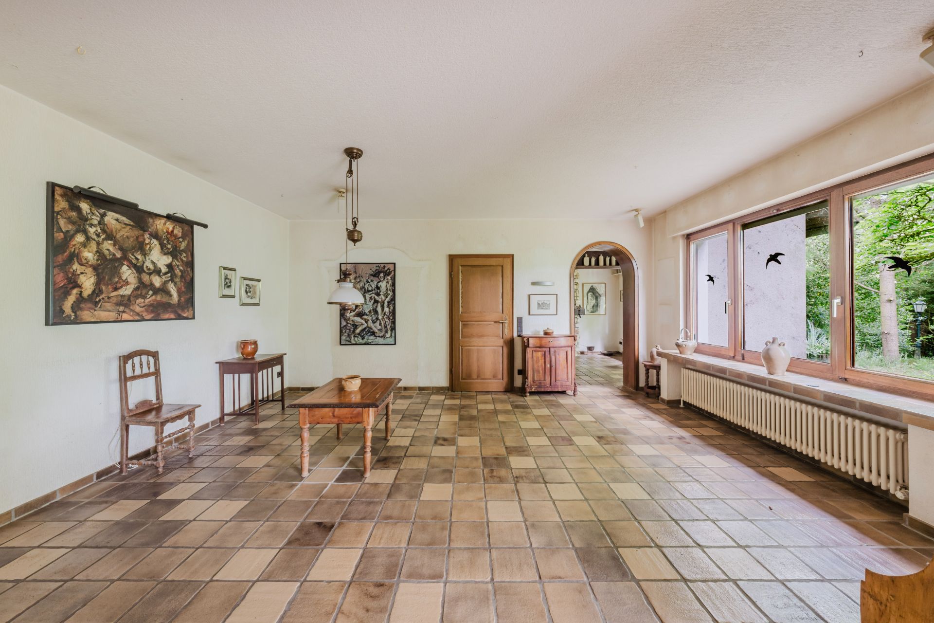Helles Wohnzimmer mit Fliesenboden, Holztisch, Stühlen, großen Fenstern und Wanddekoration.