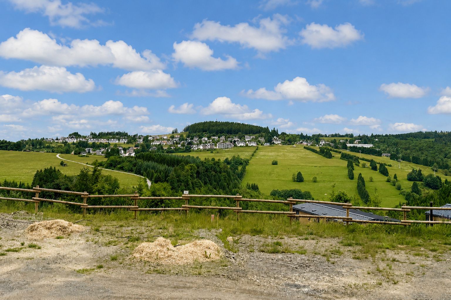 Blick auf eine grüne Hügellandschaft mit Häusern im Hintergrund und Holzzaun im Vordergrund.