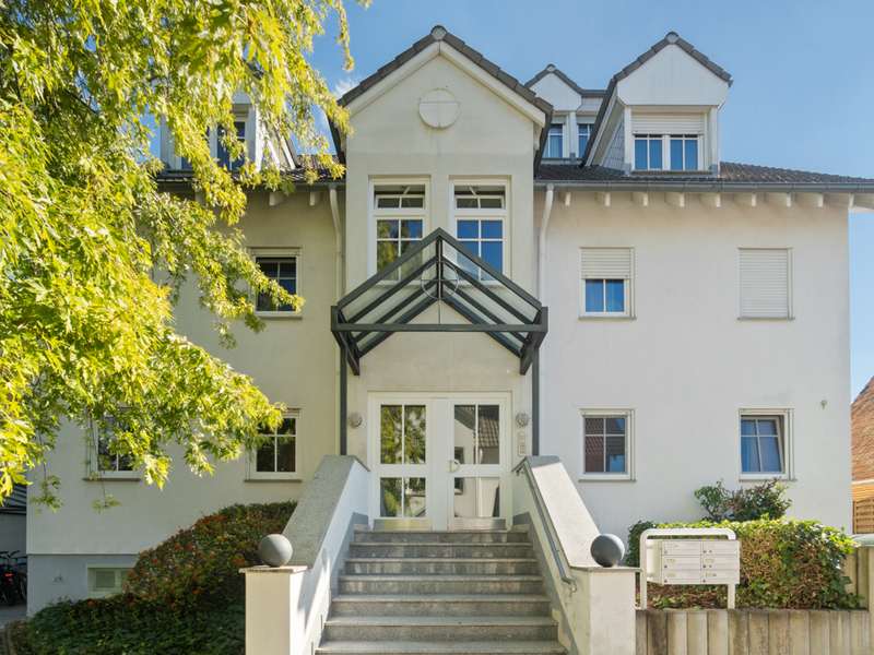 Eingang eines weißen Mehrfamilienhauses mit Treppe, Glasdach und mehreren Fenstern.