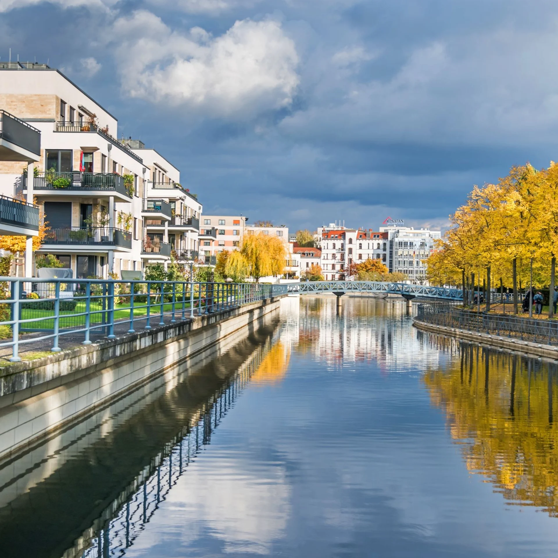Moderne Wohngebäude am Flussufer mit gelben Bäumen und Gehweg bei bewölktem Himmel.
