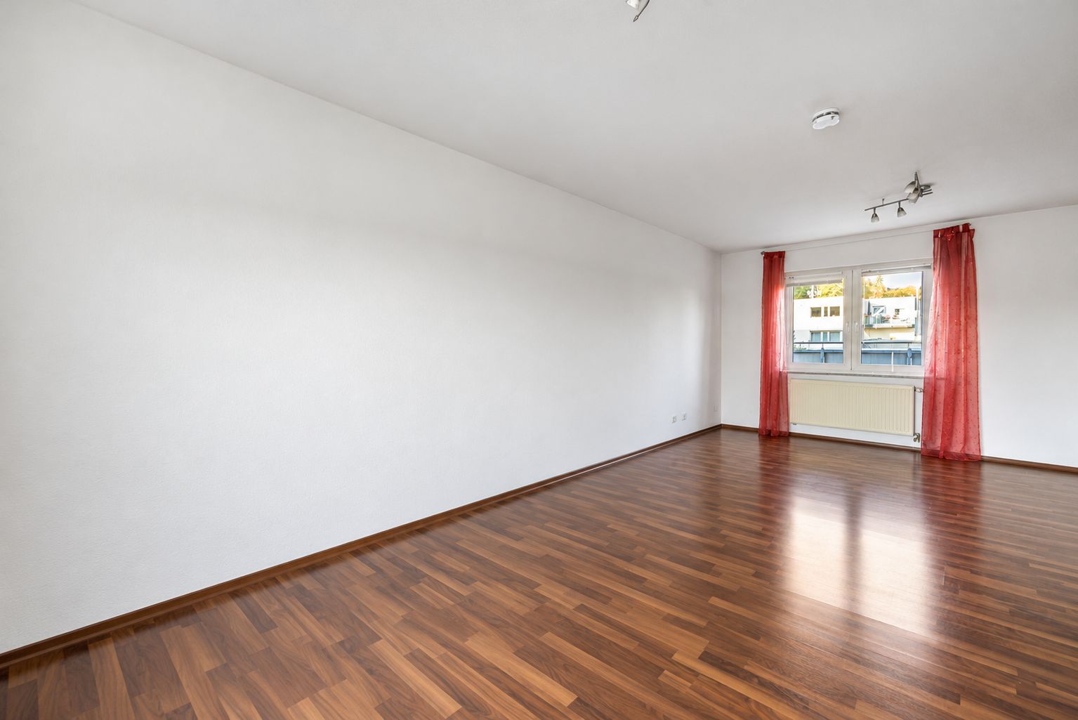 Leeres Zimmer mit Holzfußboden, weißen Wänden und Fenster mit roten Vorhängen.