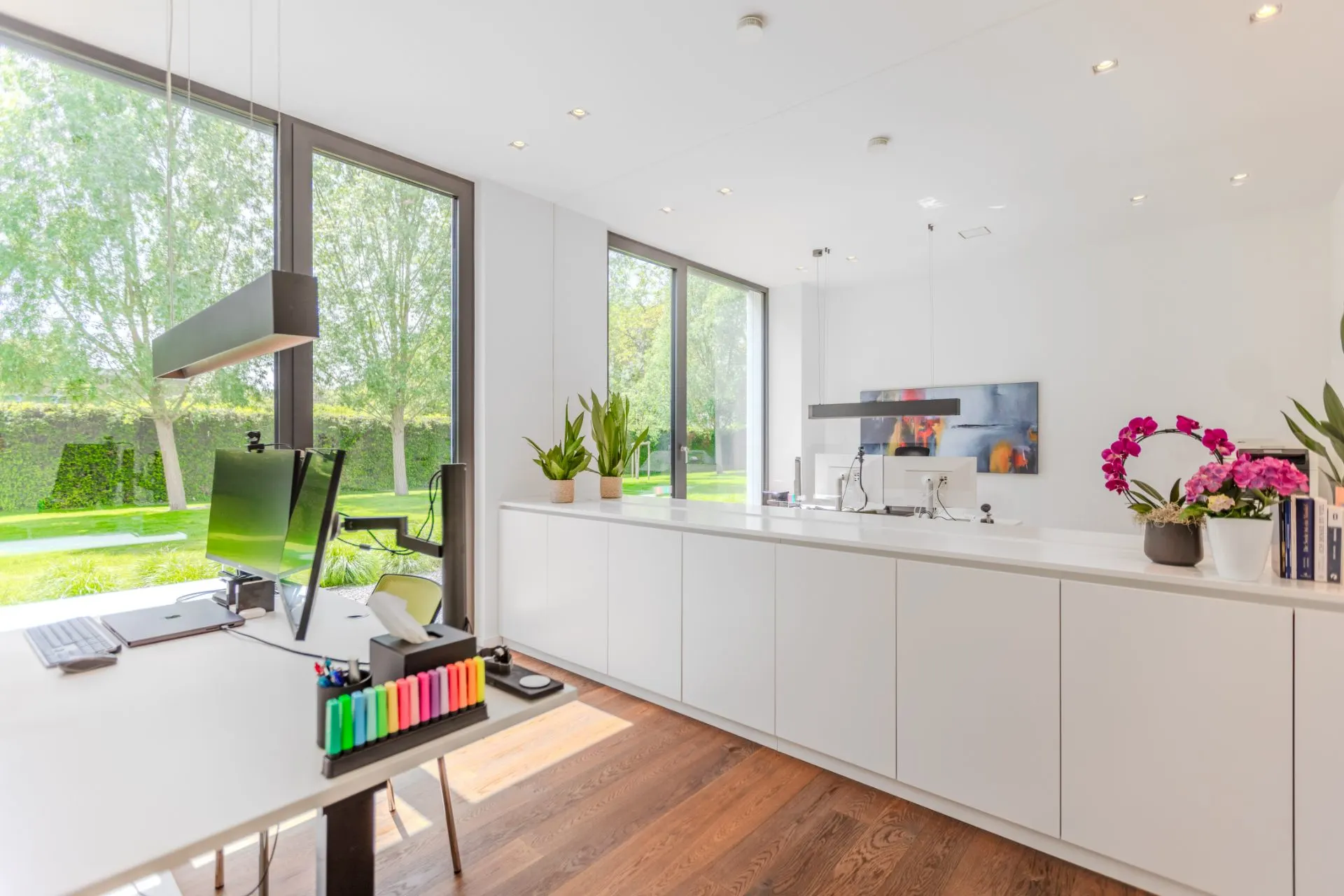 Helles modernes Arbeitszimmer mit großen Fenstern, weißen Möbeln und Pflanzen.