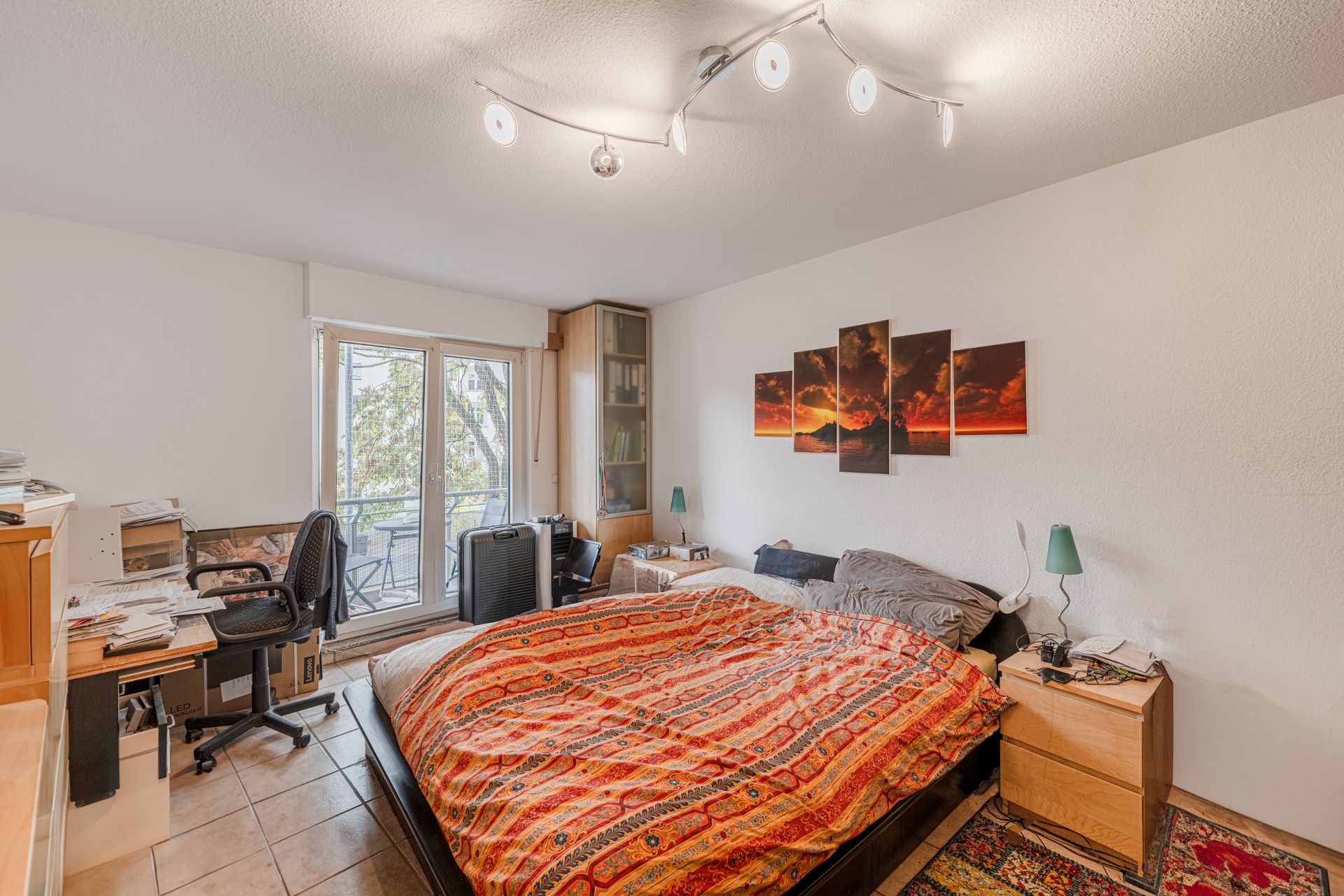 Schlafzimmer mit buntem Bett, Schreibtisch, Stuhl und Zugang zu einem Balkon.