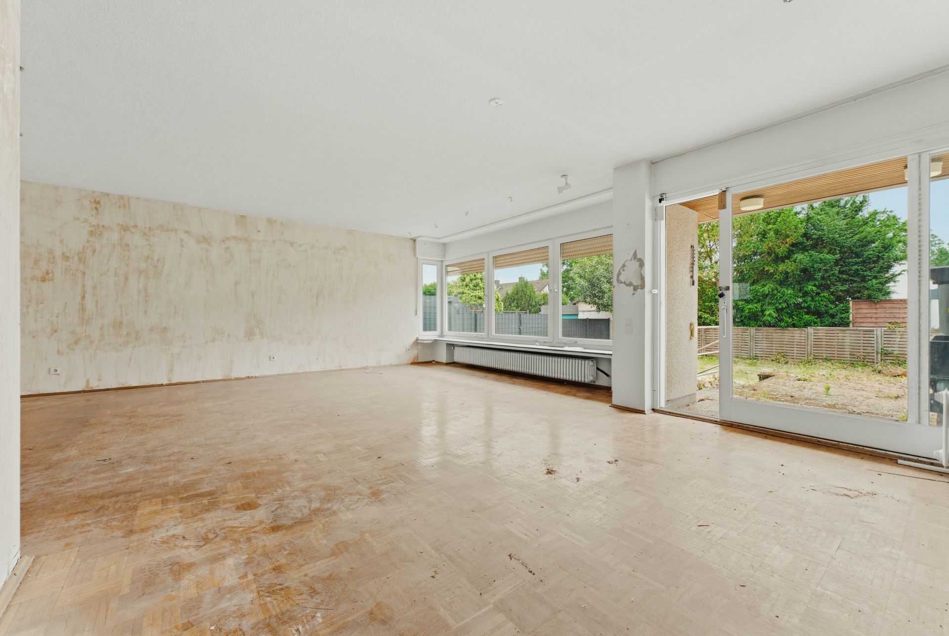 Leerer, heller Raum mit großen Fenstern, Glastür zum Garten und abgenutztem Holzfußboden.