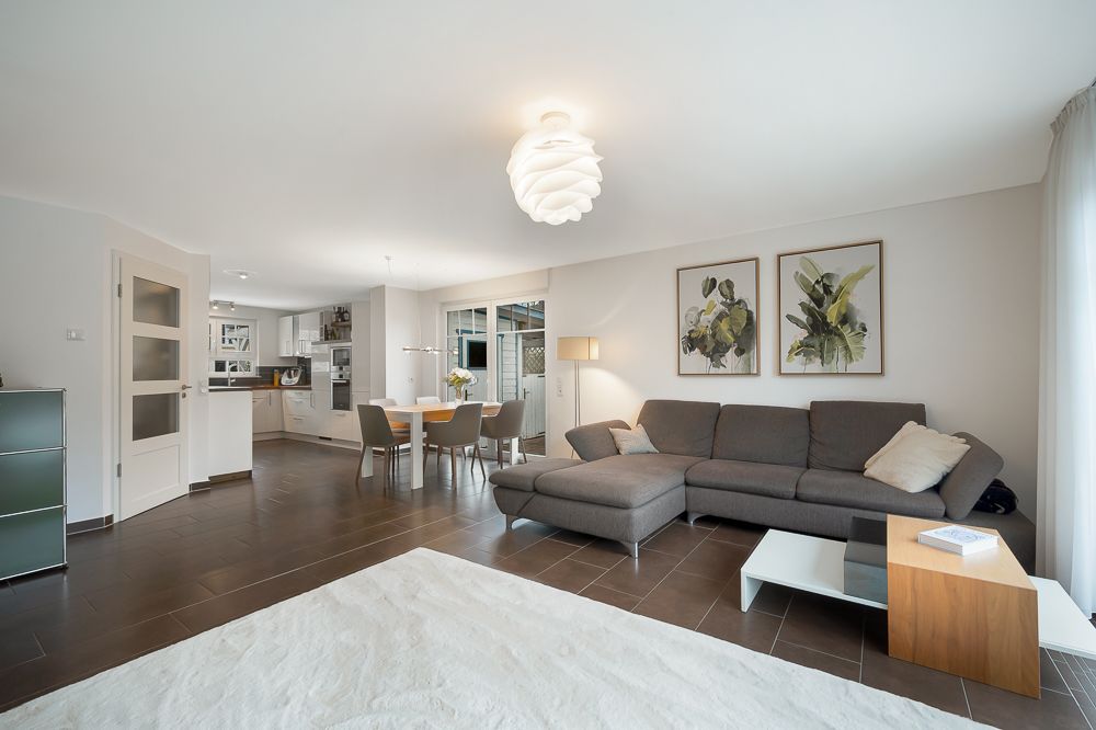Modernes Wohnzimmer mit grauem Sofa, Esstisch, offener Küche und hellem Teppich.