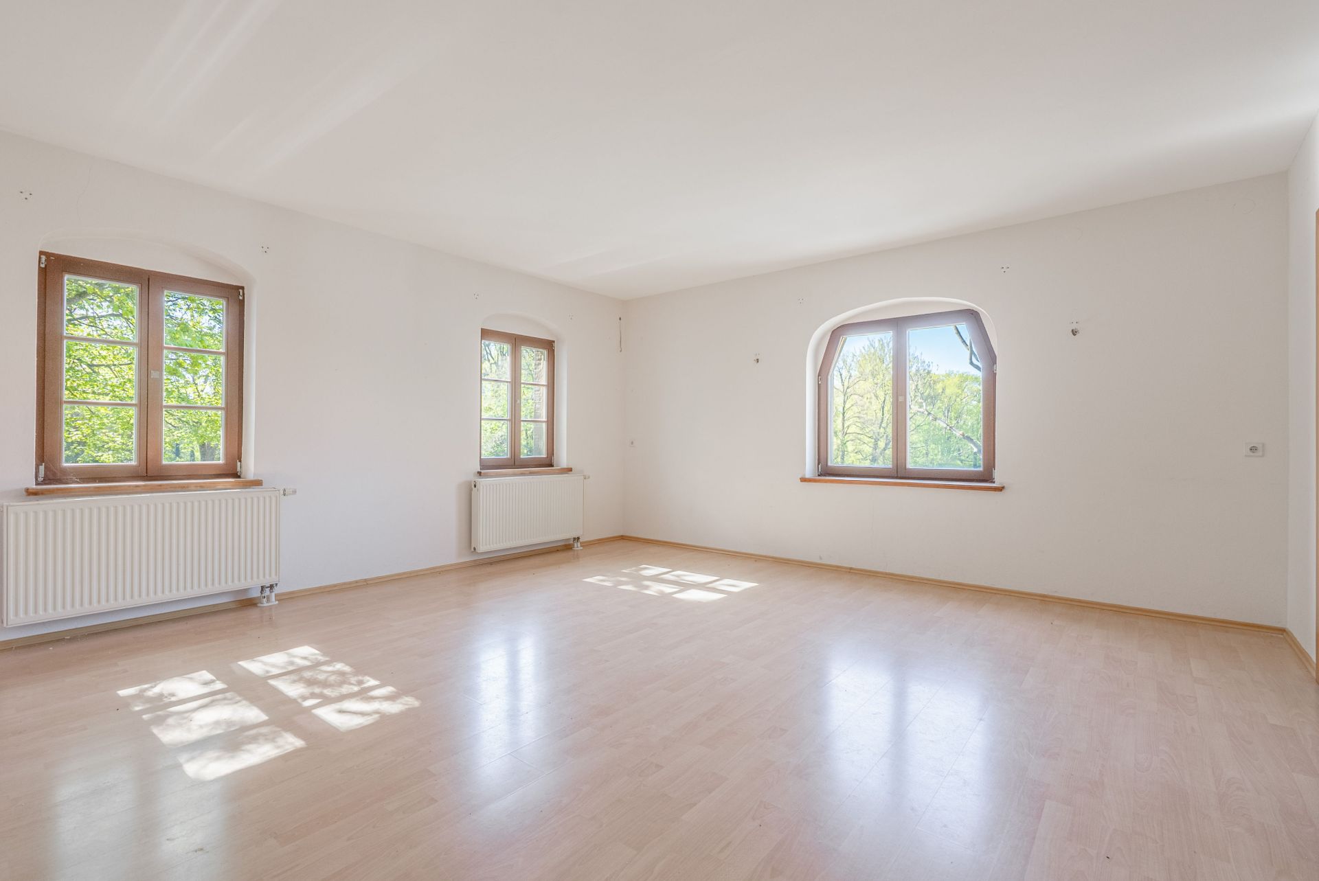 Heller, leerer Raum mit drei großen Fenstern, hellem Holzboden und weißen Wänden.