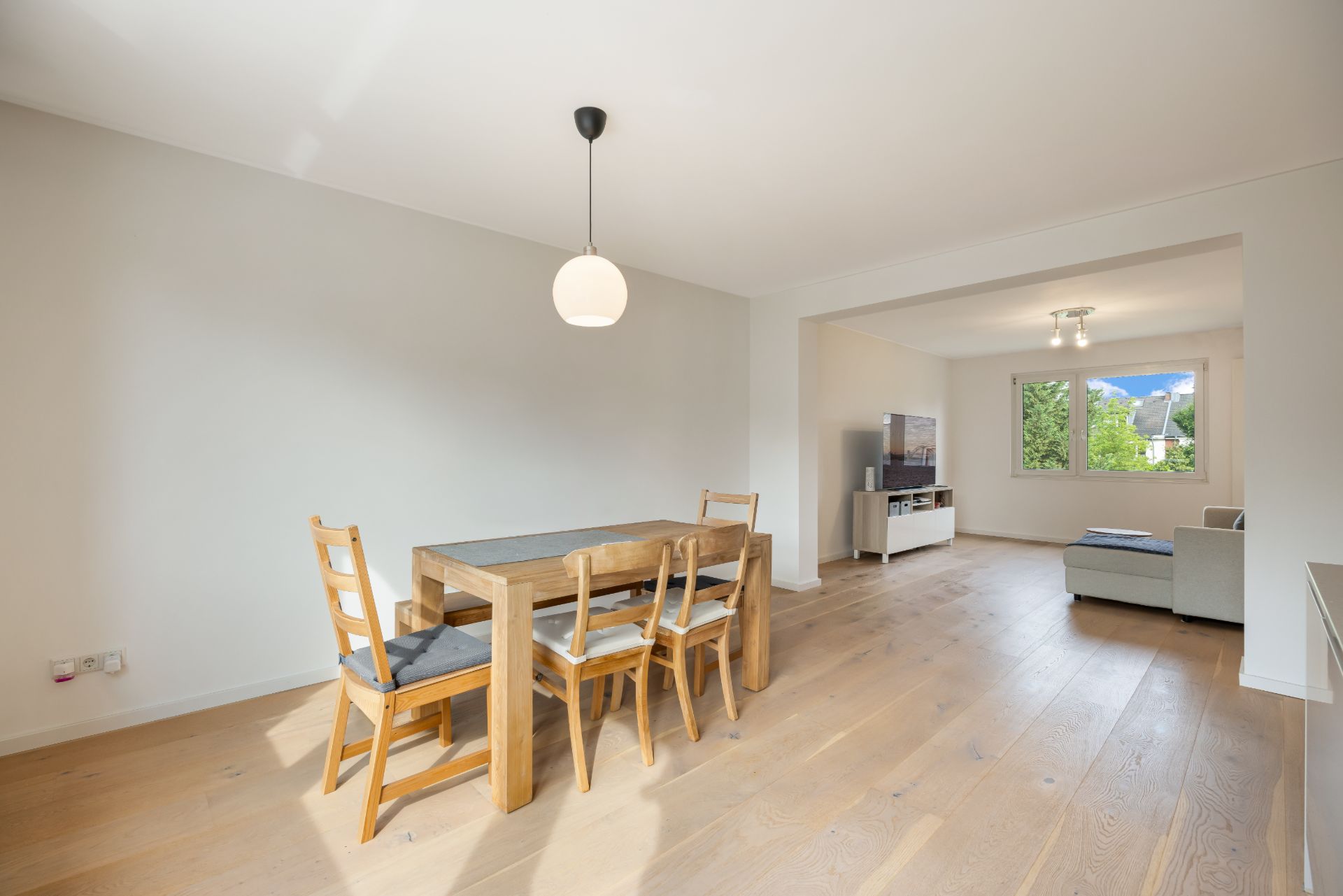 Helles Wohnzimmer mit Esstisch, vier Stühlen, Holzfußboden und Fenster mit Blick ins Grüne.