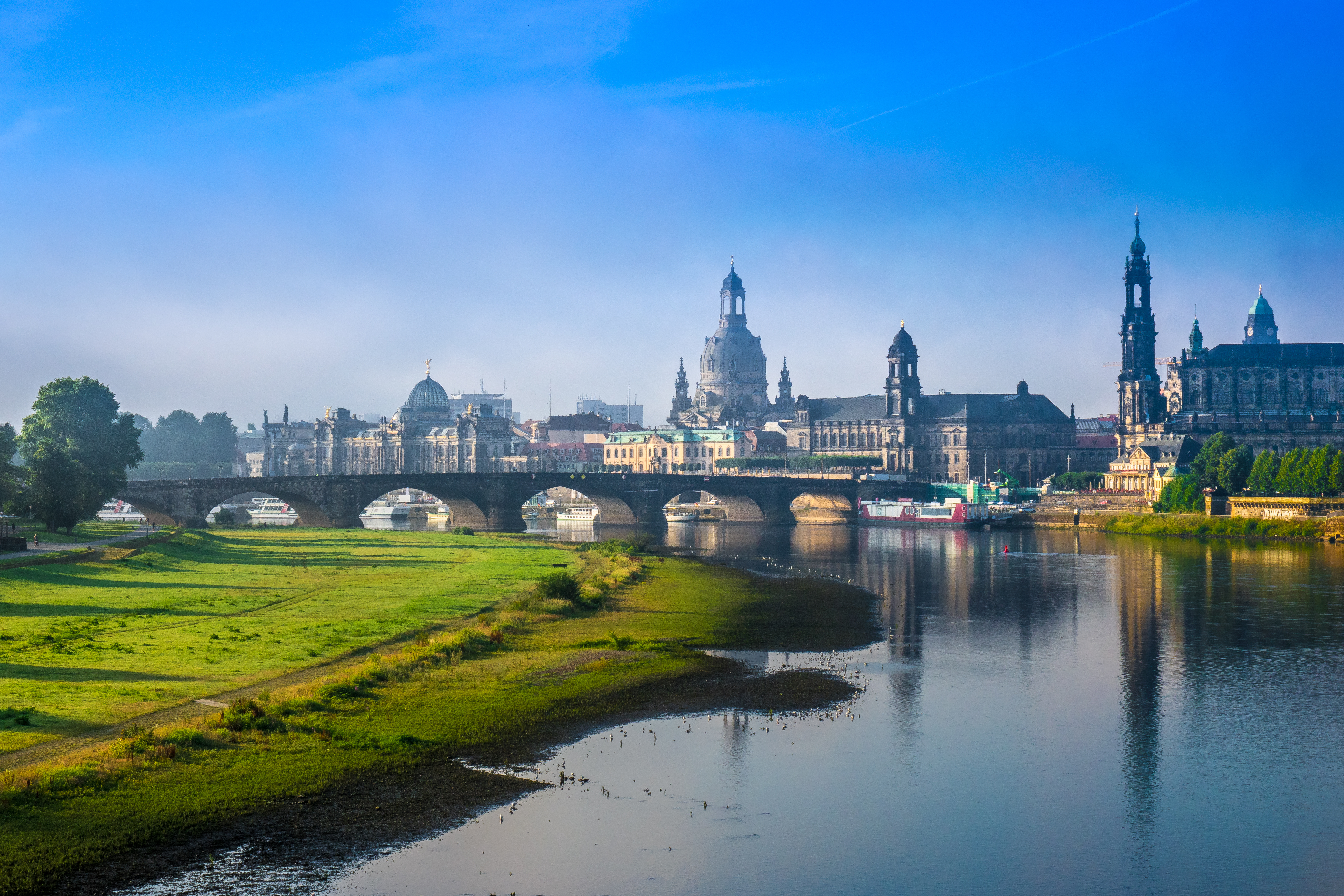 Stadtansicht mit Fluss, Brücke und historischen Gebäuden im Hintergrund.