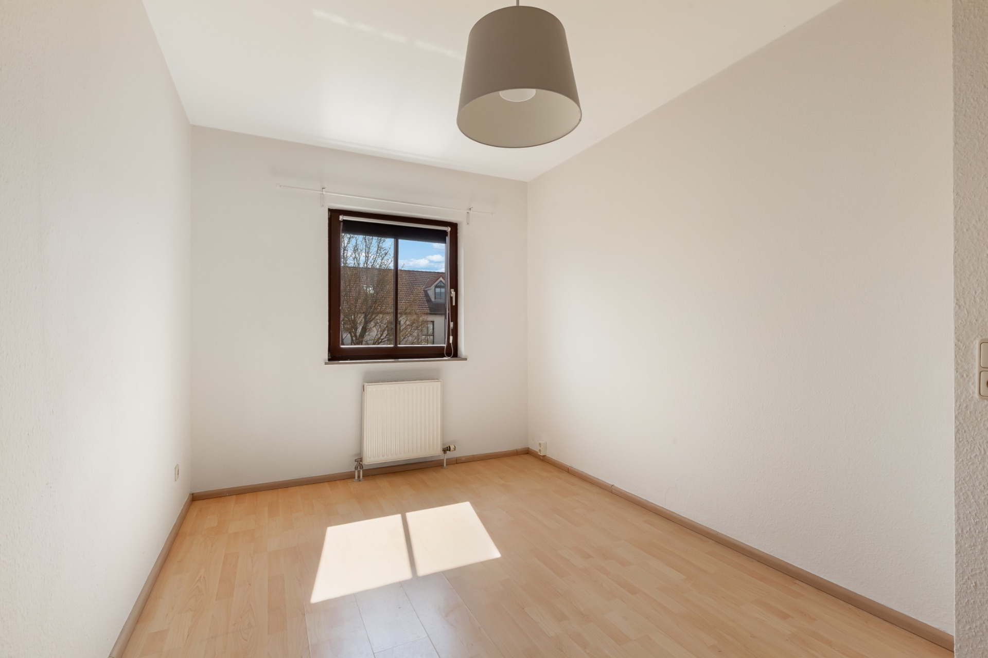 Leeres Zimmer mit Holzboden, Fenster, Heizkörper und Deckenlampe.