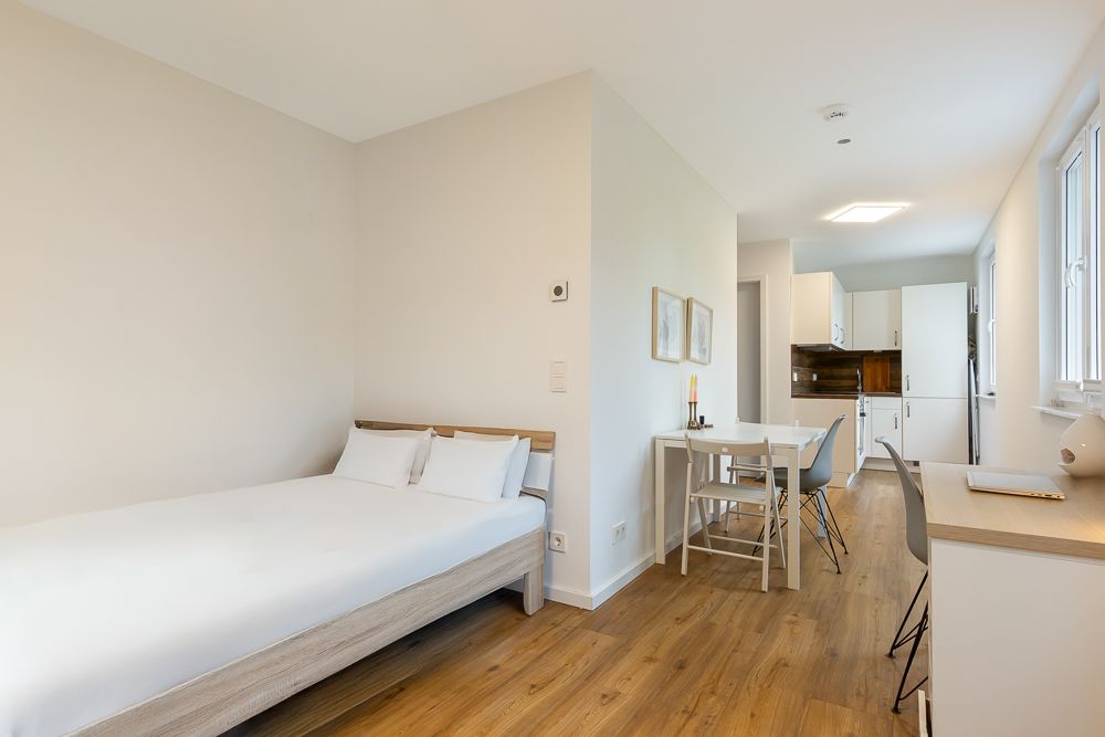 Helles Apartmentzimmer mit Bett, Schreibtisch, Esstisch und Küchenzeile auf Holzboden.
