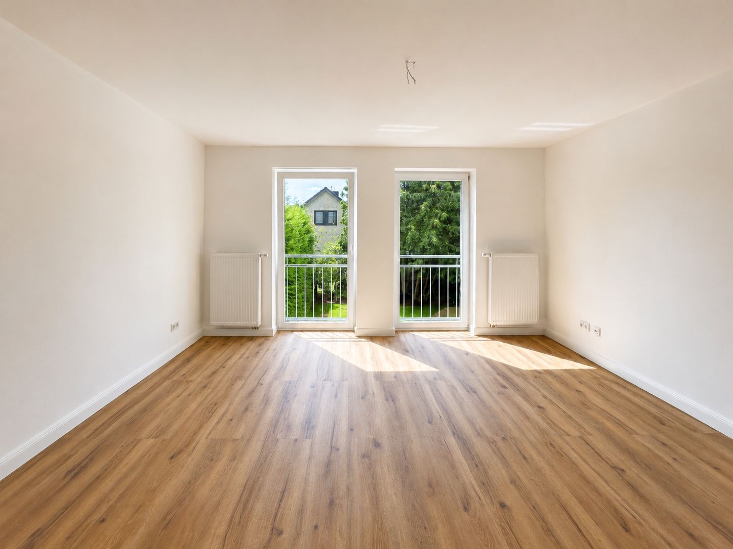 Leeres Zimmer mit Holzfußboden, zwei bodentiefen Fenstern und weißen Wänden.