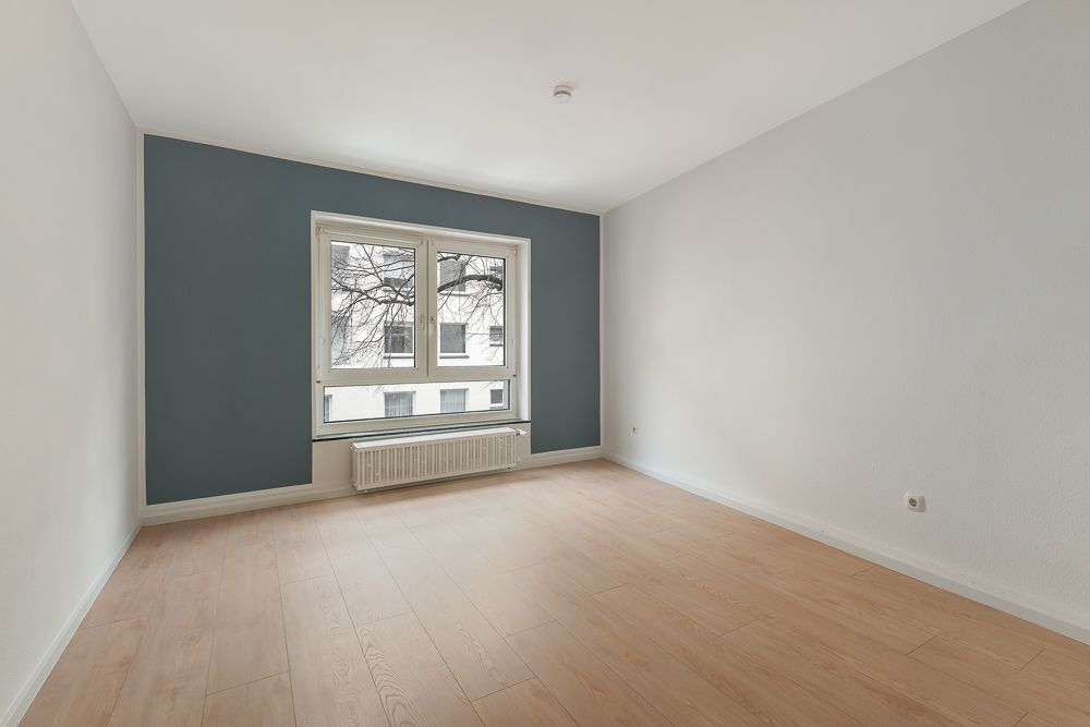 Leeres Zimmer mit Holzfußboden, blau-grauer Wand und großem Fenster mit Heizkörper.