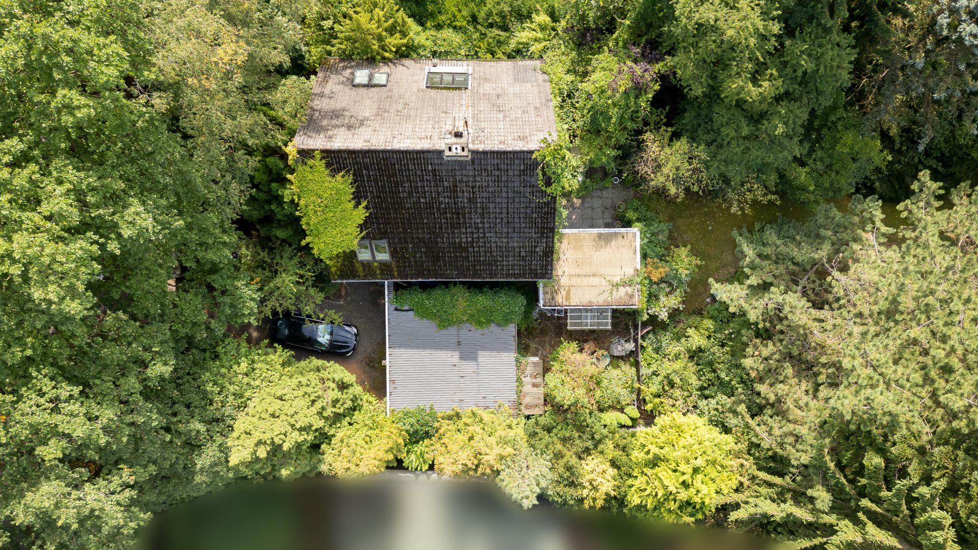 Luftaufnahme eines Hauses mit dunklem Dach, umgeben von vielen Bäumen und Grünflächen.