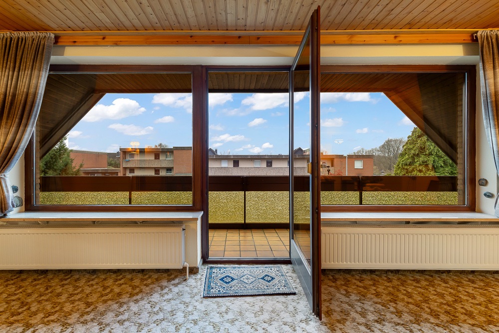 Wohnzimmer mit Teppichboden und offener Tür zum Balkon mit Blick auf Nachbargebäude.