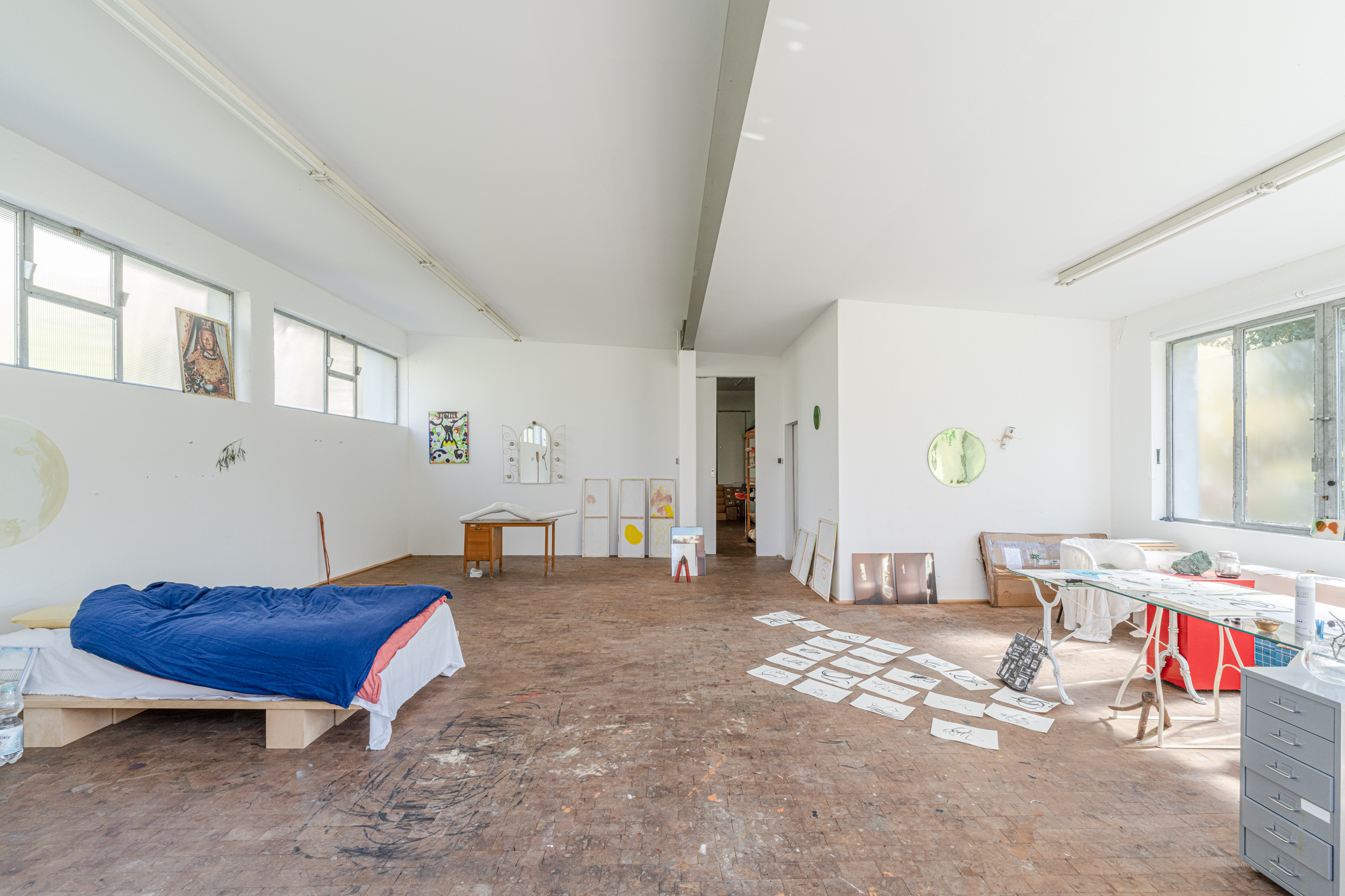 Helles Zimmer mit Bett, Schreibtisch, Zeichnungen auf dem Boden und großem Fenster.