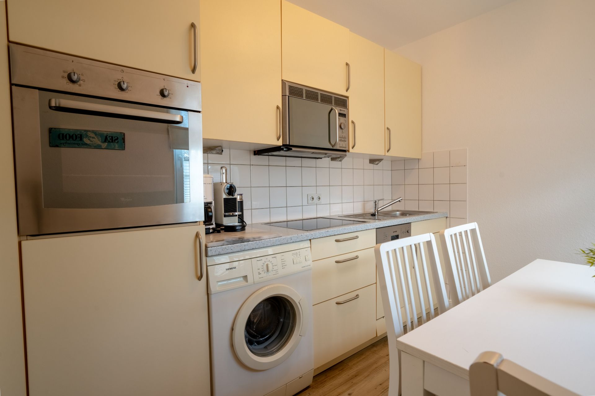 Kleine Küche mit Waschmaschine, Backofen, Mikrowelle und Esstisch mit zwei Stühlen.