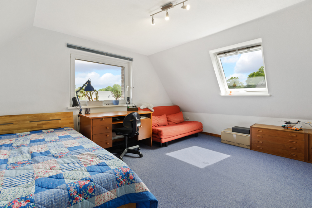 Helles Schlafzimmer mit blauem Teppich, Bett, Schreibtisch, rotem Sofa und zwei Fenstern.