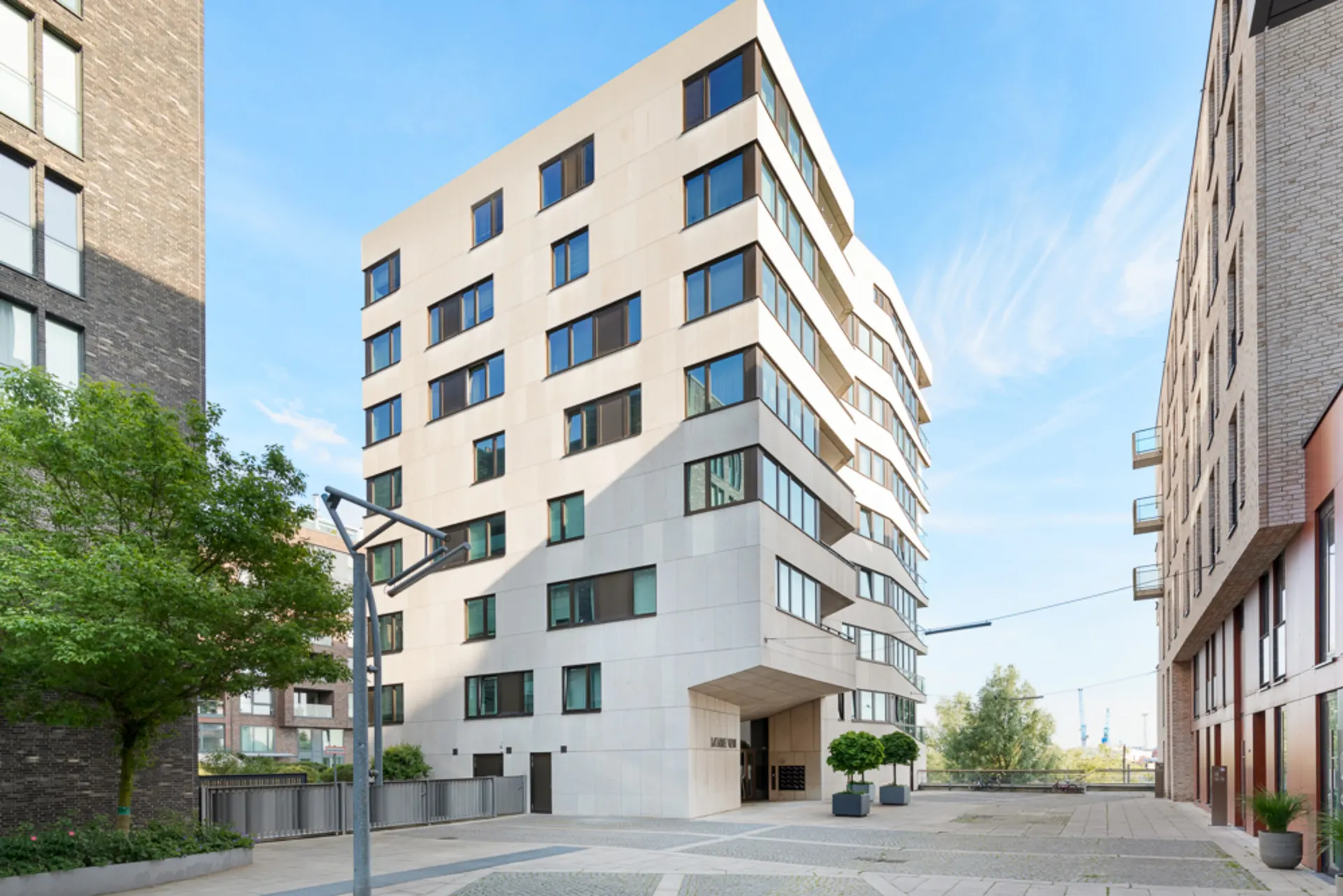 Modernes Wohngebäude mit vielen Fenstern zwischen zwei anderen Gebäuden, vorne Bäume und ein Platz.