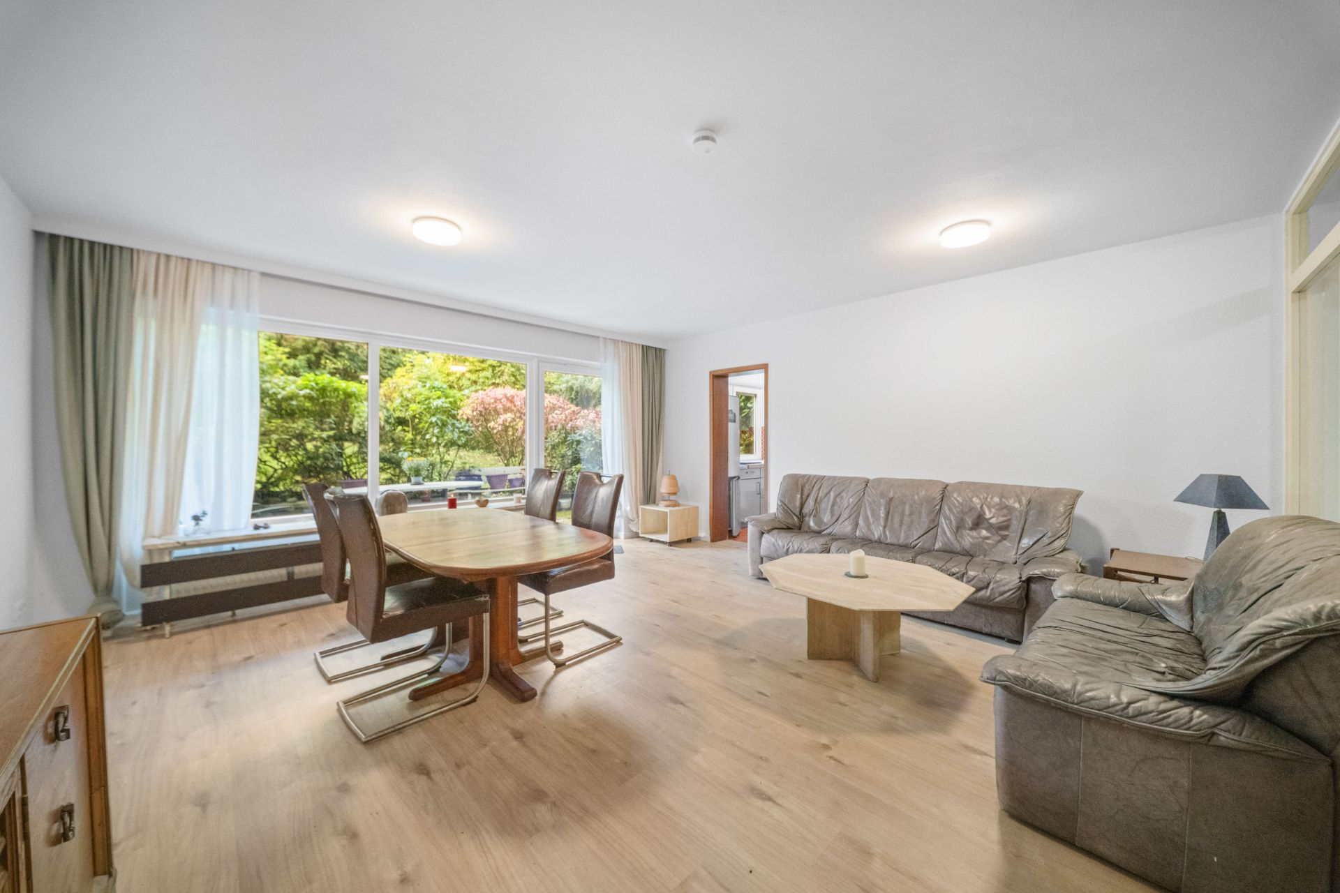 Helles Wohnzimmer mit Esstisch, Sofas, Couchtisch und großen Fenstern mit Blick auf einen Garten.