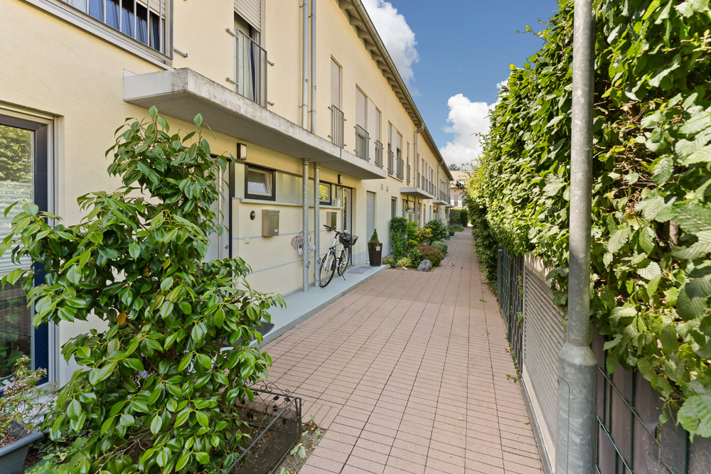 Gepflasterter Gehweg vor Reihenhäusern mit Fahrrädern und Pflanzen am Eingang.