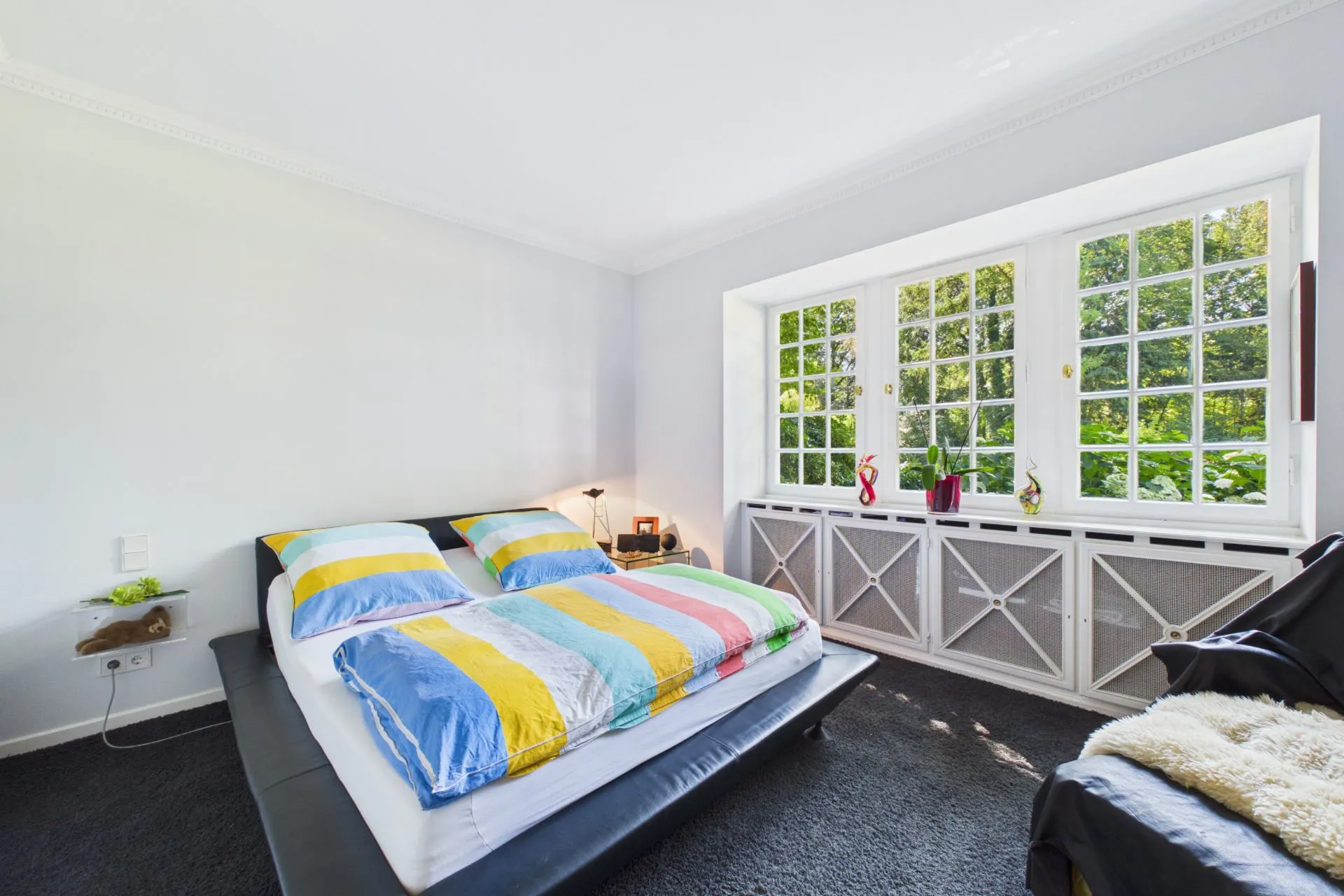 Helles Schlafzimmer mit Doppelbett, buntem Bettbezug und großem Fenster mit Blick ins Grüne.