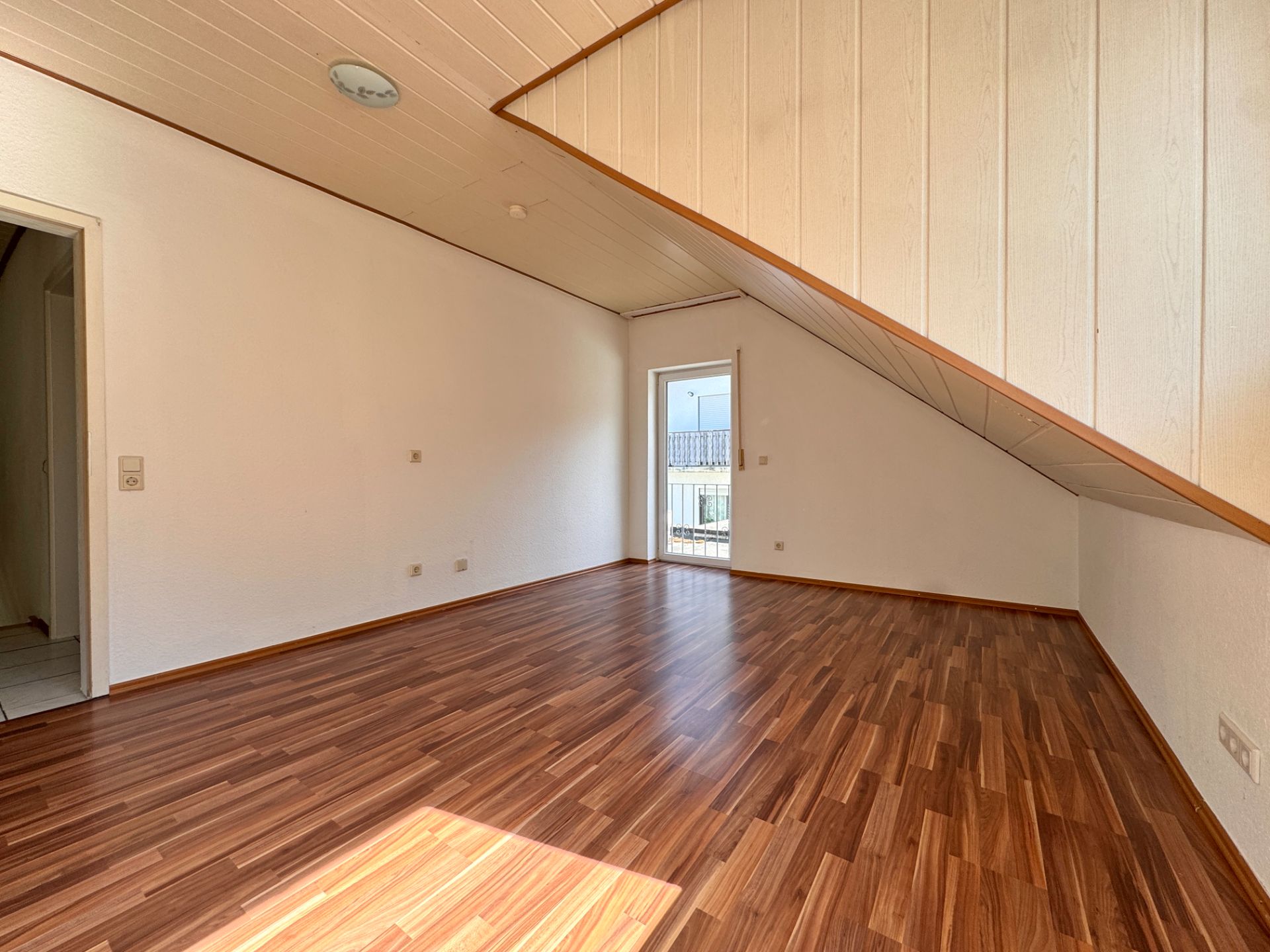 Helles Zimmer mit Holzfußboden, Dachschräge und Zugang zu einem Balkon.