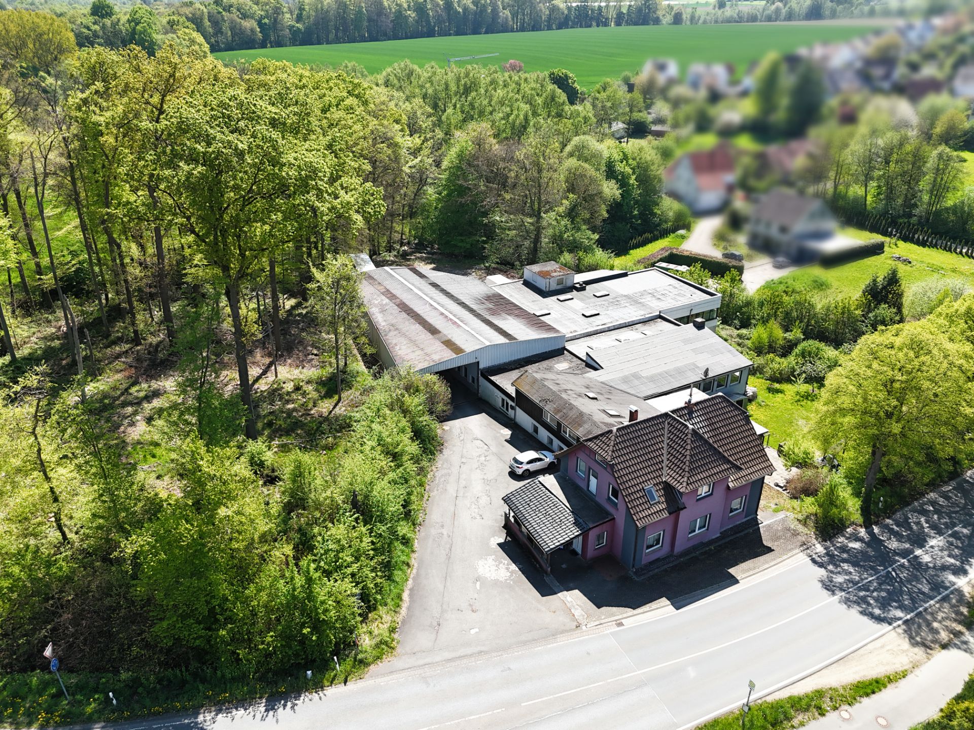 Wohnhaus mit Nebengebäuden, umgeben von Bäumen und einer Landstraße im Grünen.