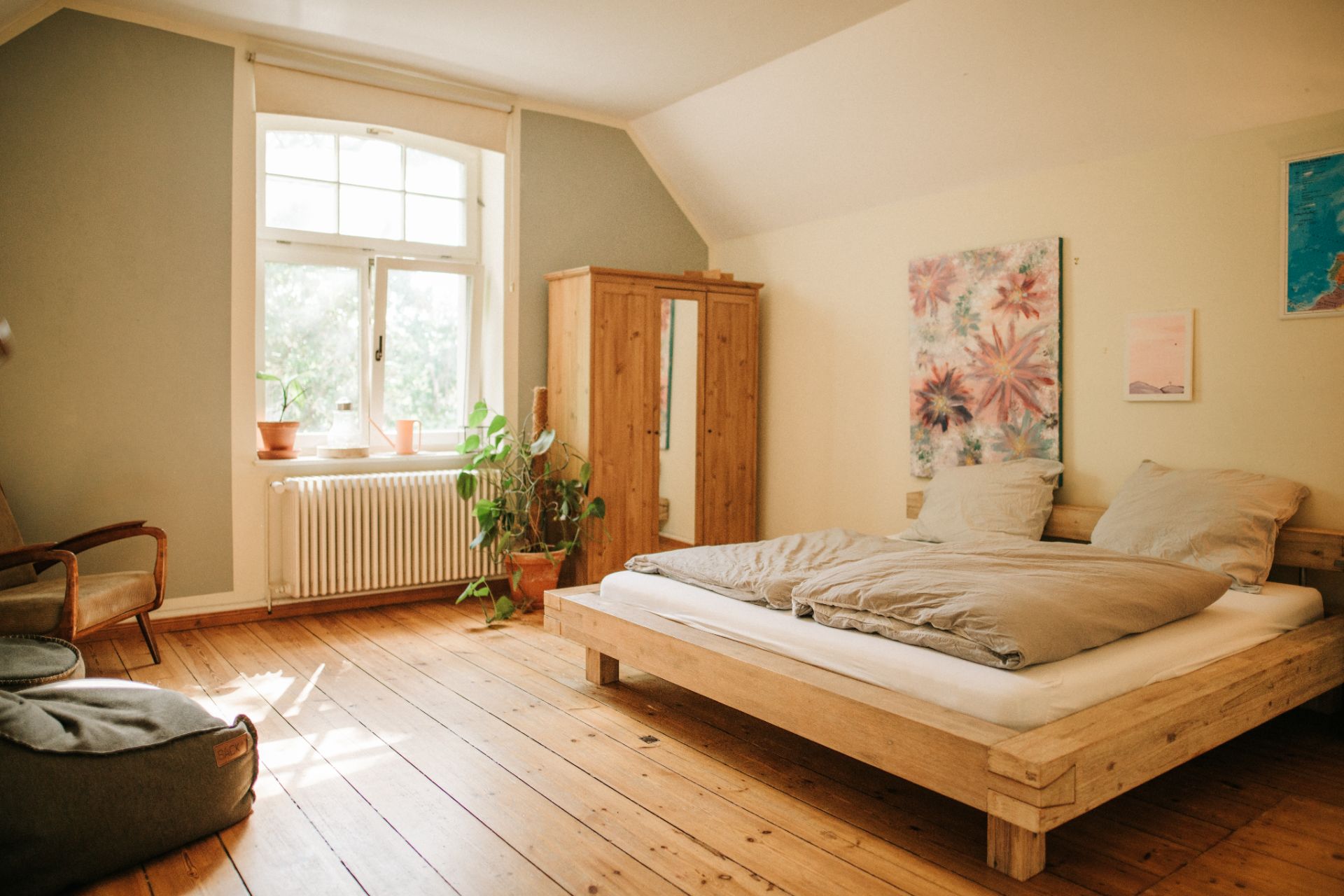Helles Schlafzimmer mit Holzboden, großem Bett, Schrank, Fenster und Pflanzen.