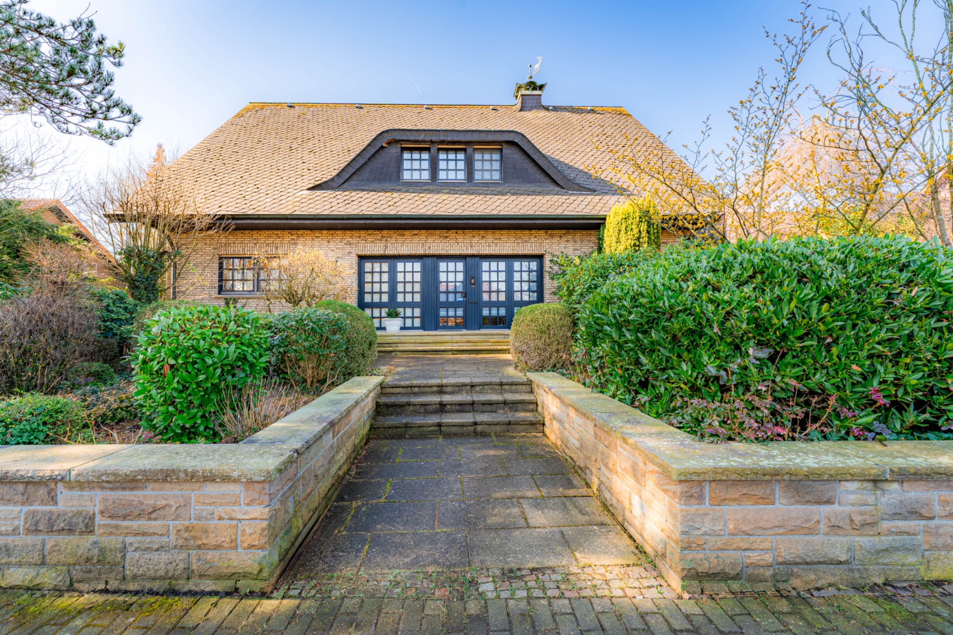 Gemütliches Haus mit großem Schrägdach, breitem Eingang und Garten mit Sträuchern.