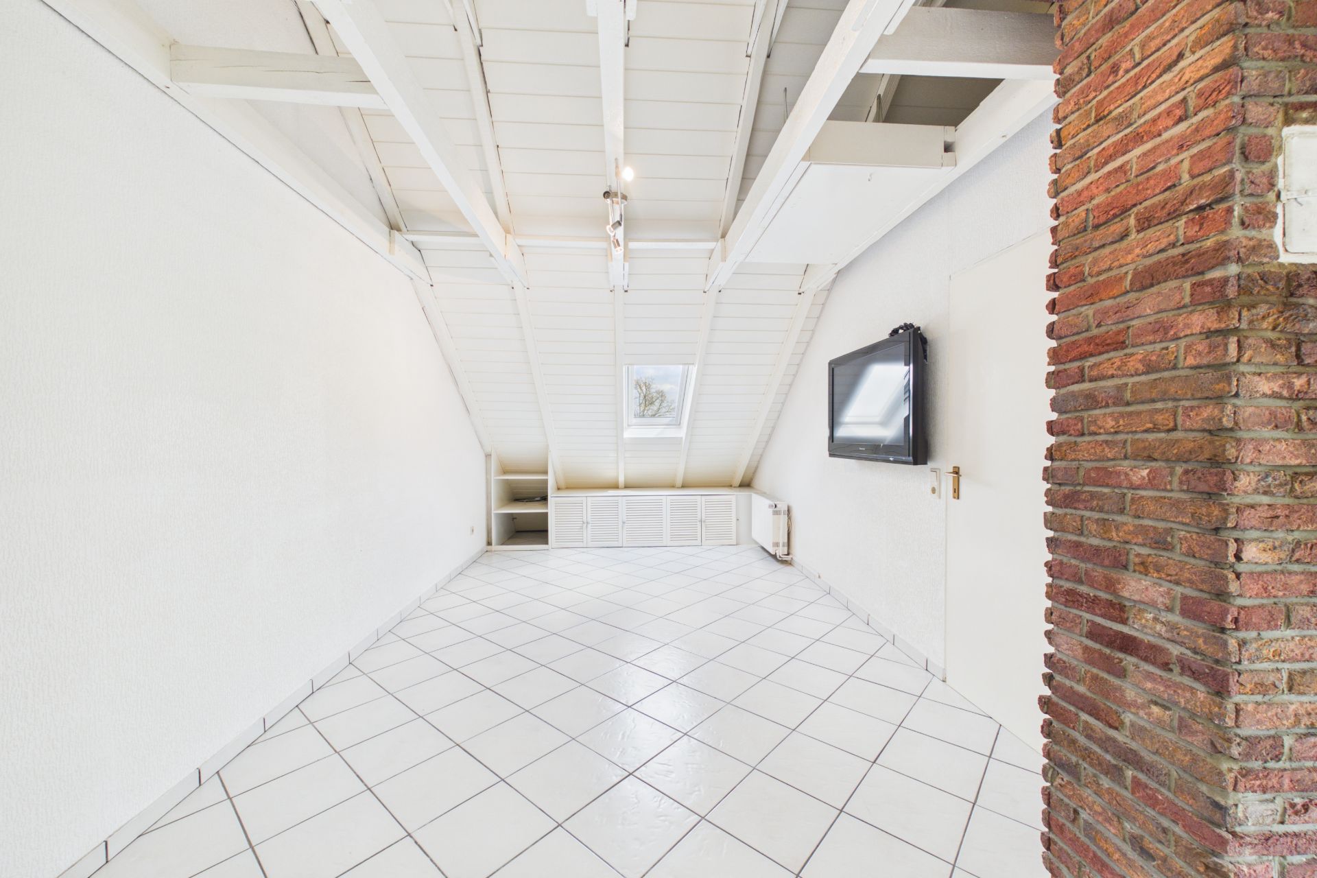 Heller Dachraum mit weißen Fliesen, schräger Decke und einem Fernseher an der Wand.