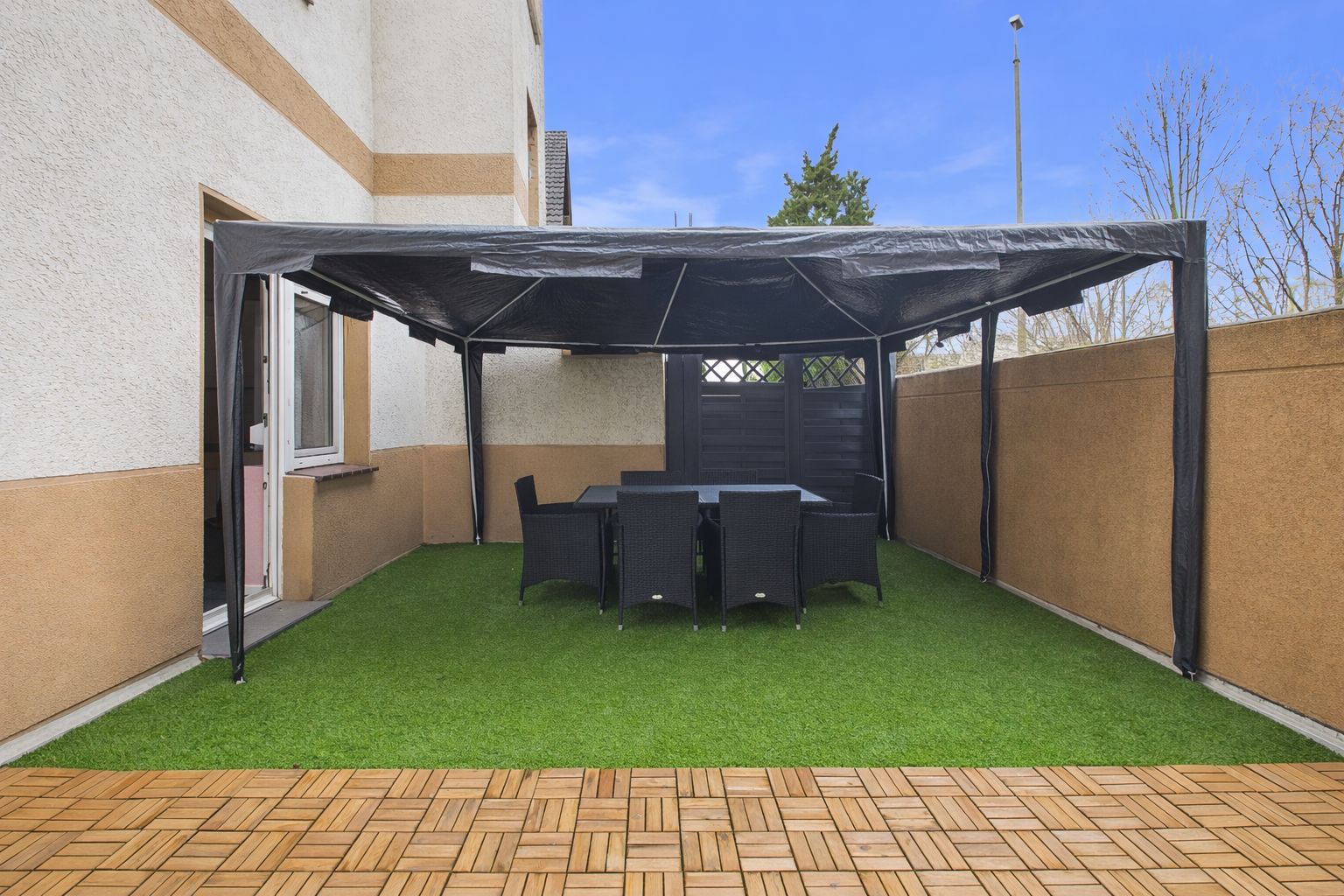 Überdachte Terrasse mit Esstisch und Stühlen auf Kunstrasen neben einem Wohnhaus.