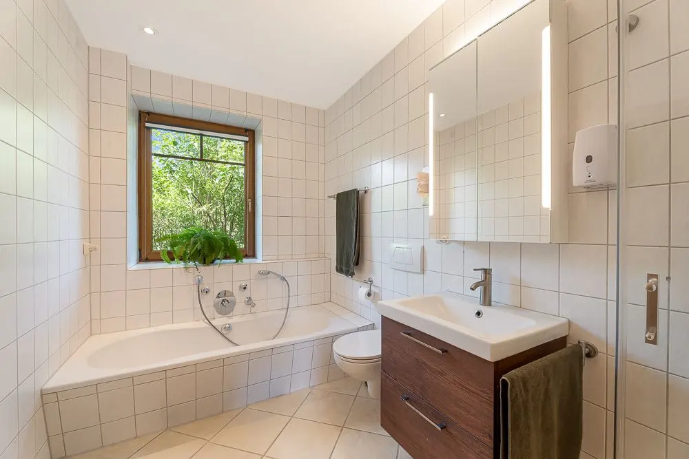 Helles Badezimmer mit Badewanne, Waschbecken, Spiegel, Fenster und grüner Pflanze auf der Fensterbank.