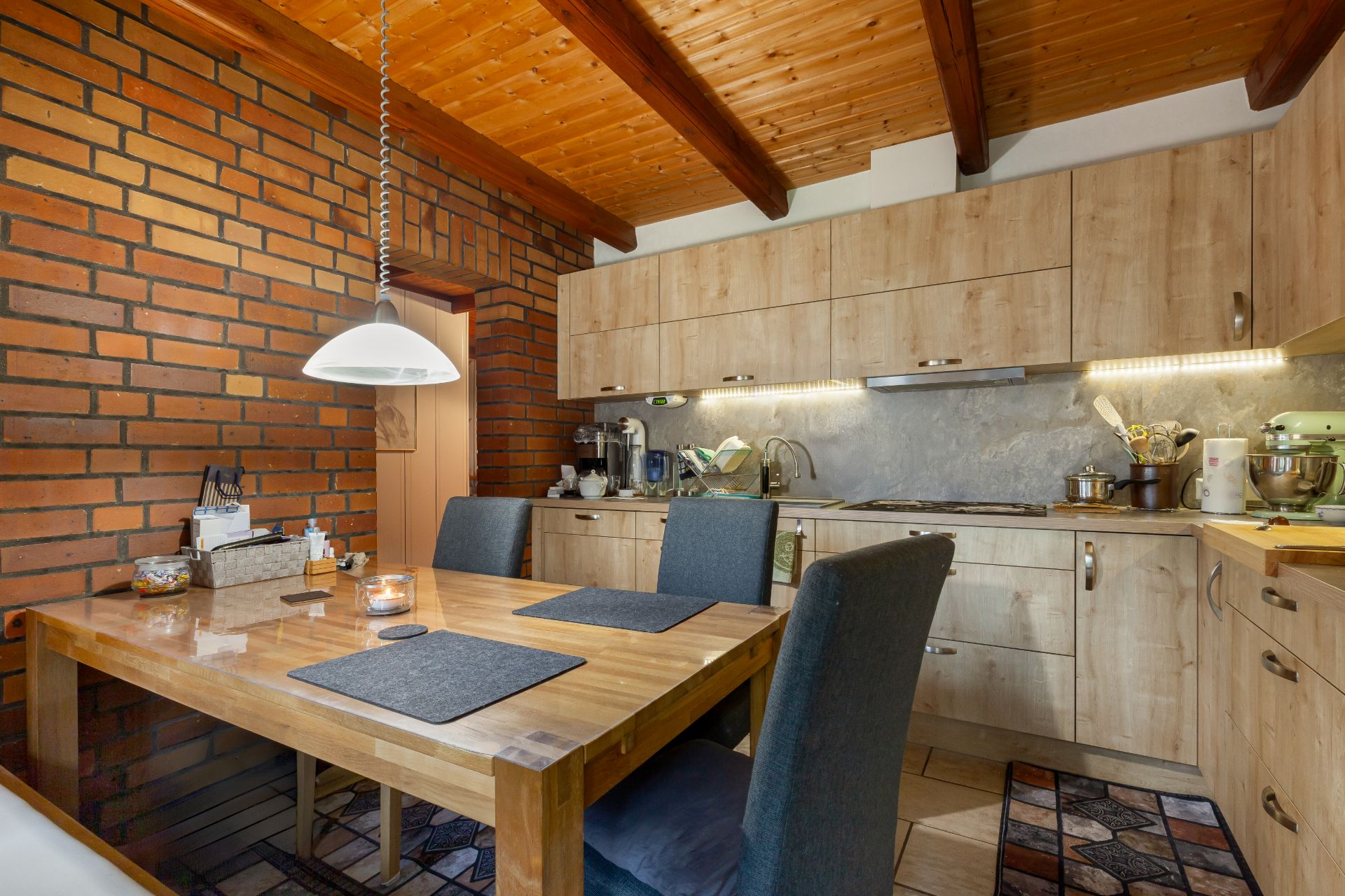 Gemütliche Küche mit Holztisch, vier Stühlen, rustikaler Backsteinwand und hölzernem Küchenschrank.