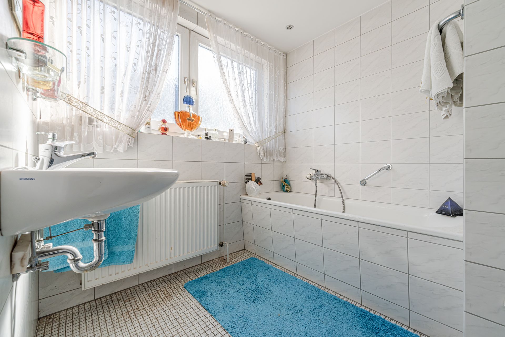 Helles Badezimmer mit Badewanne, Waschbecken, blauem Teppich und Fenster mit Gardinen.