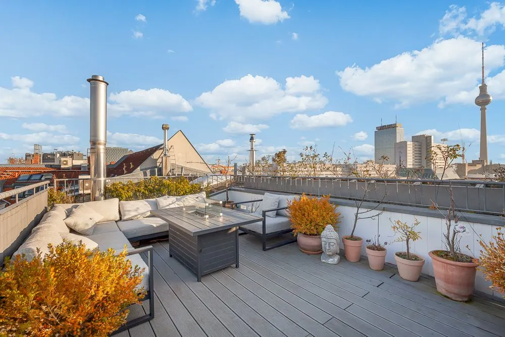 Dachterrasse mit Sitzgruppe, Tisch und mehreren Pflanzentöpfen, Blick auf Stadtgebäude.