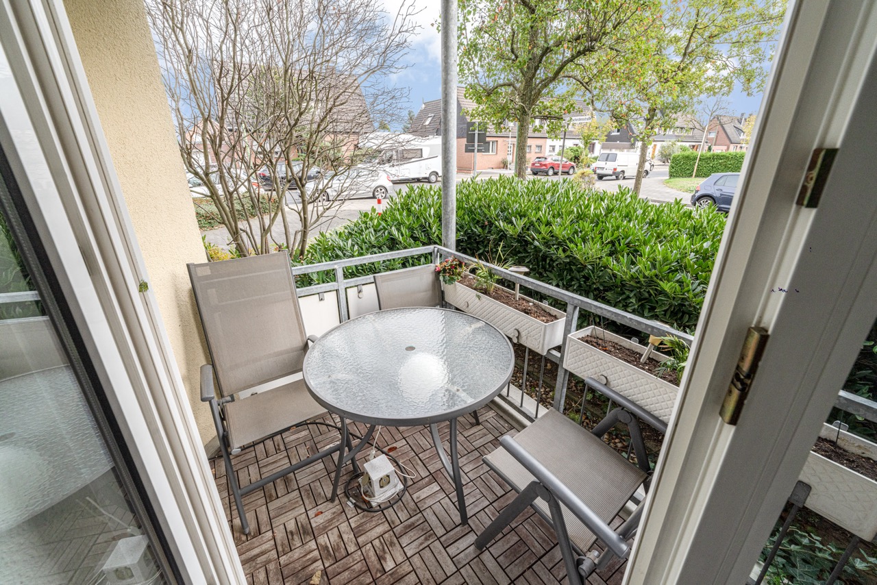 Balkon mit rundem Glastisch, vier Stühlen und Blumenkästen, Blick auf Straße und Bäume.