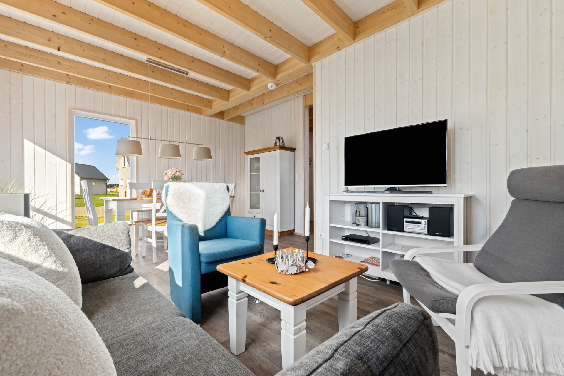 Helles Wohnzimmer mit Sofa, Sesseln, Couchtisch, Fernseher und sichtbaren Holzbalken an der Decke.