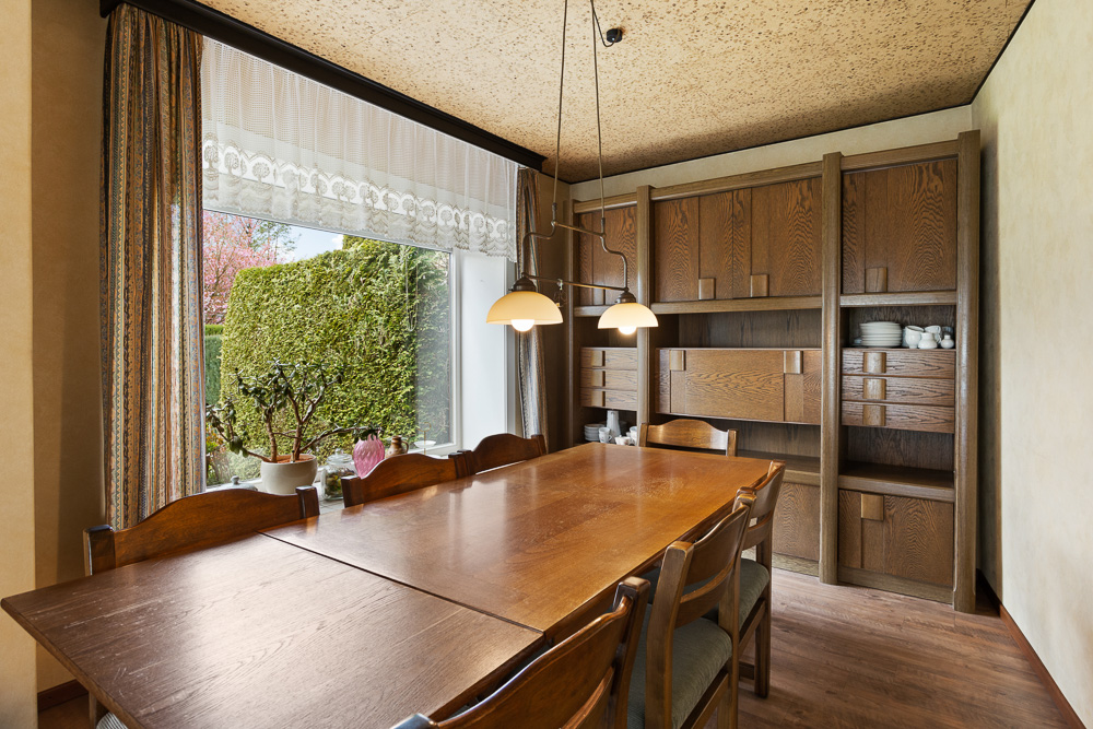 Esszimmer mit großem Holztisch, Stühlen und Holzregal, daneben großes Fenster mit Blick ins Grüne.